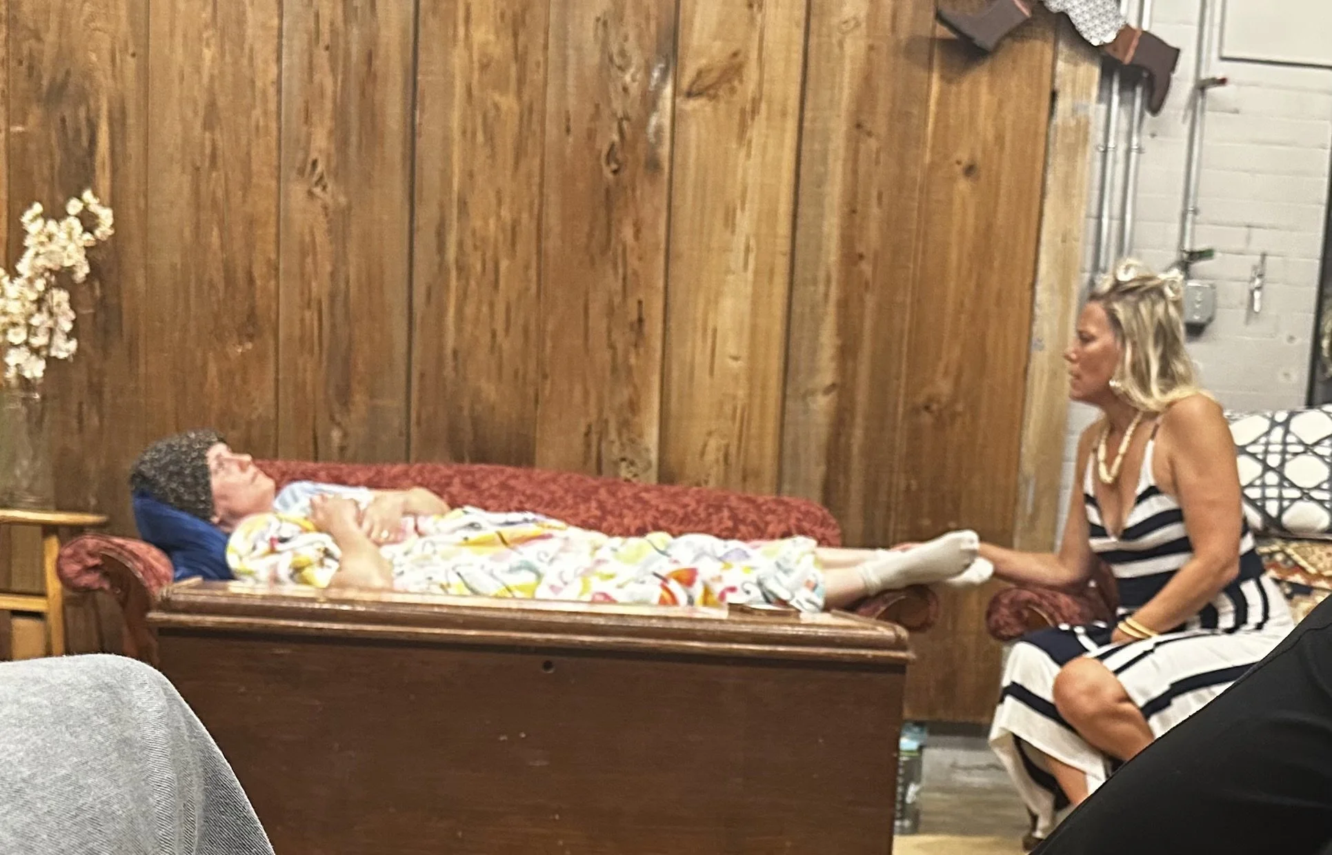 A woman dressed in a black and white striped dress is sitting by a vintage chaise lounge, holding hands with an elderly woman lying down on the chaise lounge. The elderly woman is wearing a colorful top and a curly wig, with her eyes closed and hands