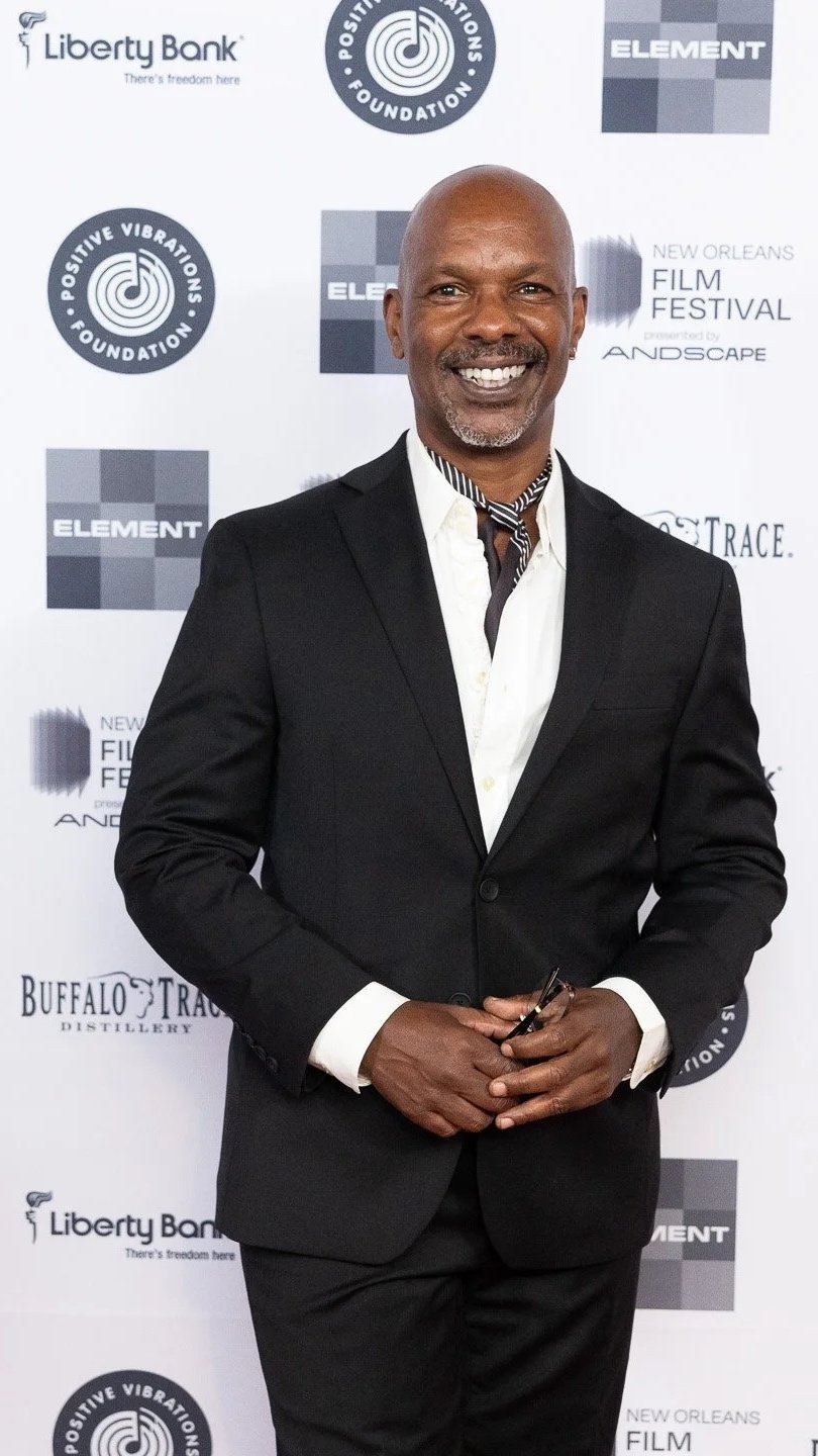 A smiling man in a black suit and white shirt standing in front of a backdrop with event logos, holding glasses in his hands.