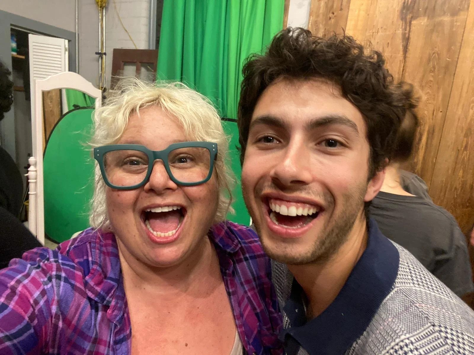 A woman with curly blonde hair and large blue glasses smiling widely beside a young man with dark curly hair, both taking a selfie in a room with a green screen, wooden walls, and other people in the background.