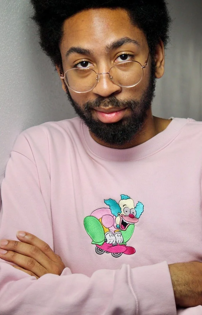 A young man with glasses and a beard wearing a pink sweatshirt featuring a cartoon clown on a skateboard.