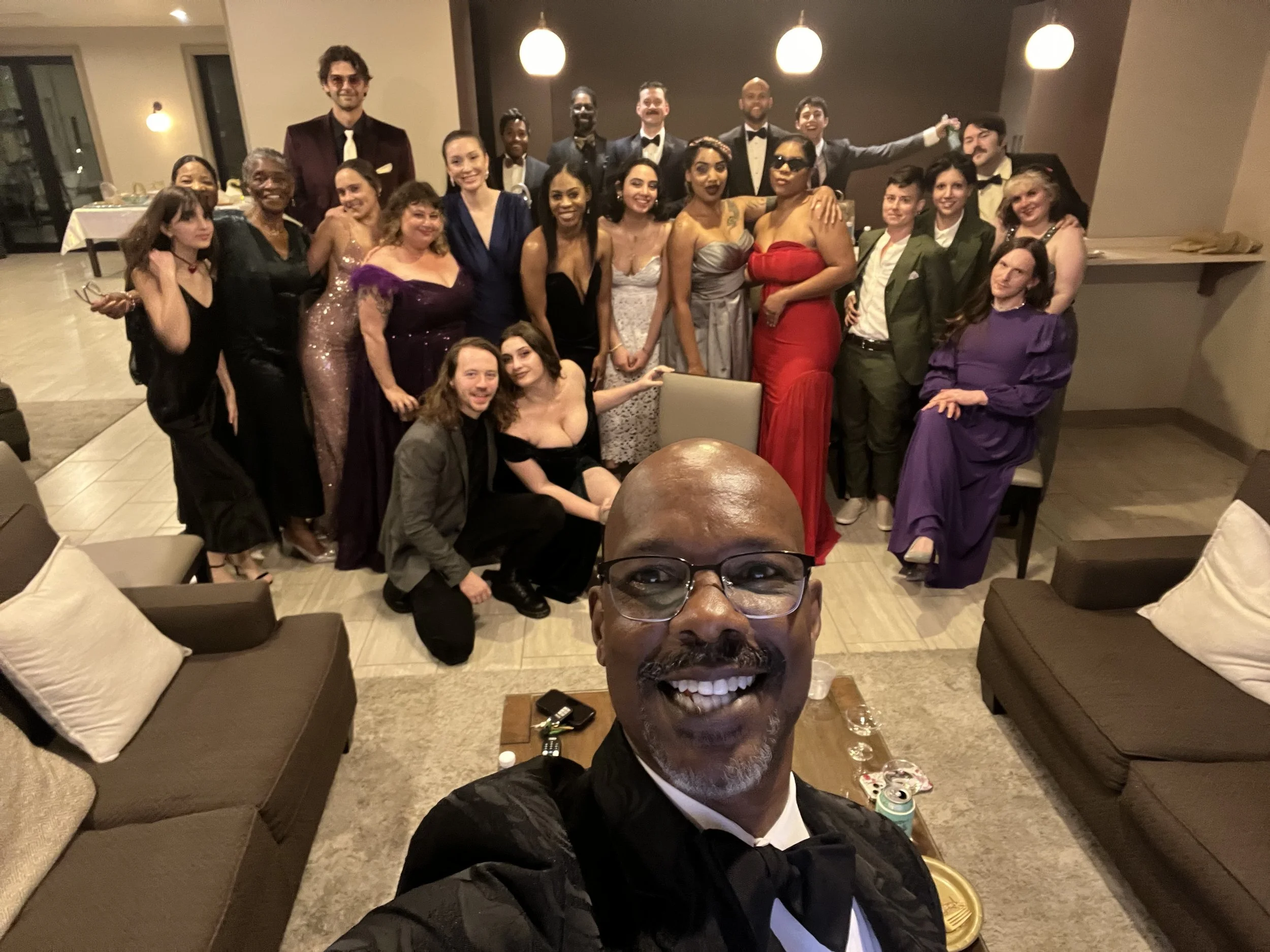 A group of people dressed in formal attire posing for a photo at a social event or party in a well-lit indoor setting, with some seated on sofas and others standing behind them.