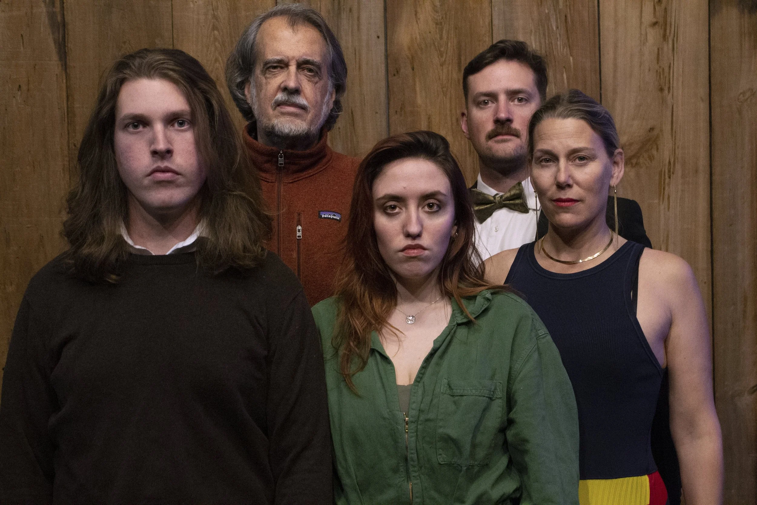 Group of five people standing in front of wooden wall, with serious expressions.