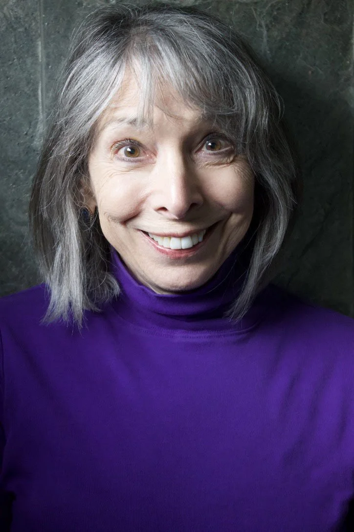 A woman with gray hair wearing a purple turtleneck, smiling and standing against a dark background.