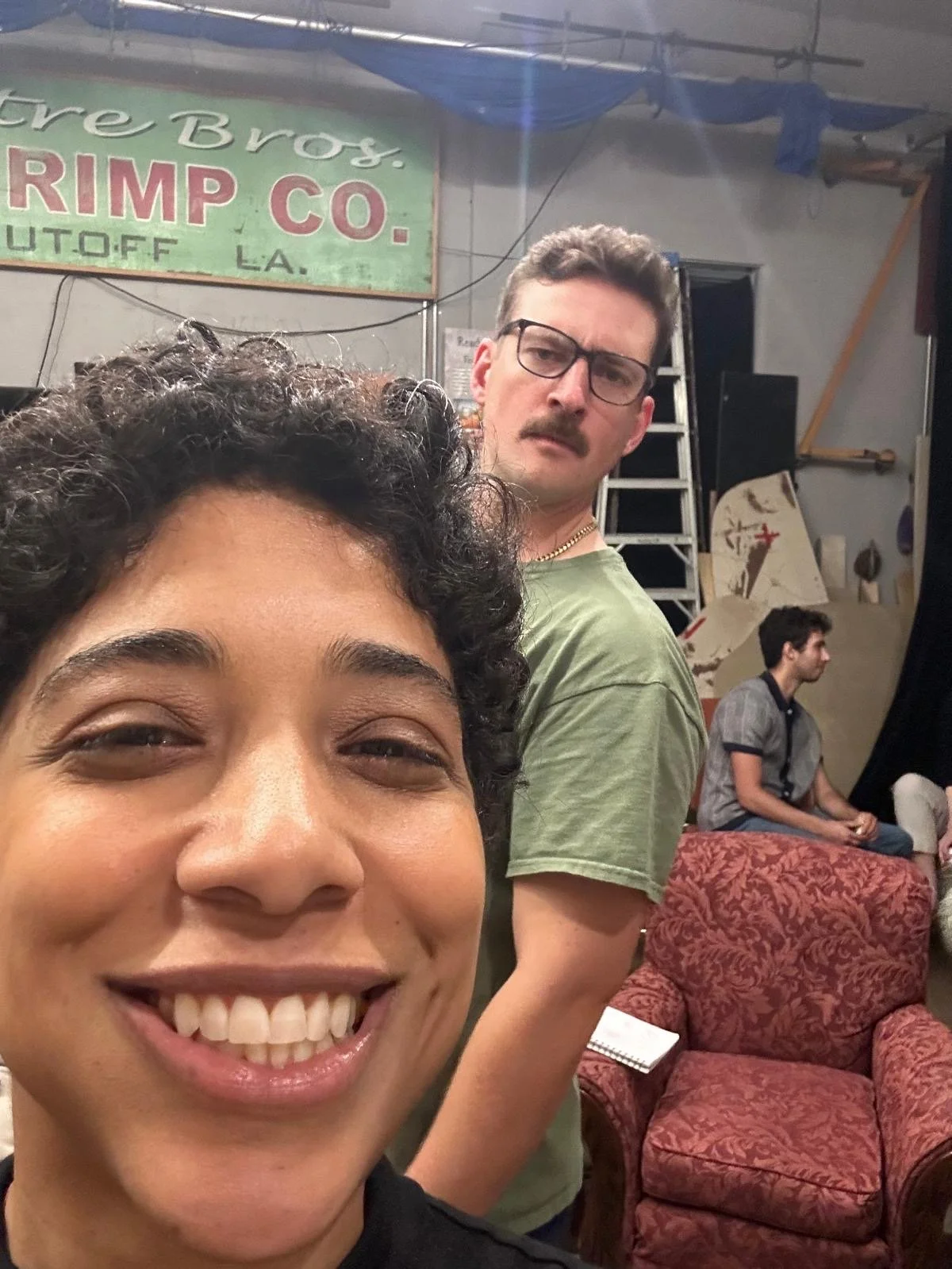 A woman smiling at the camera with two men in the background, one wearing glasses and a green shirt, the other sitting on a red patterned armchair. Behind them is a sign that reads 'Friere Bros. Shrimp Co. Outfitter LA' and a ladder, with a cluttered