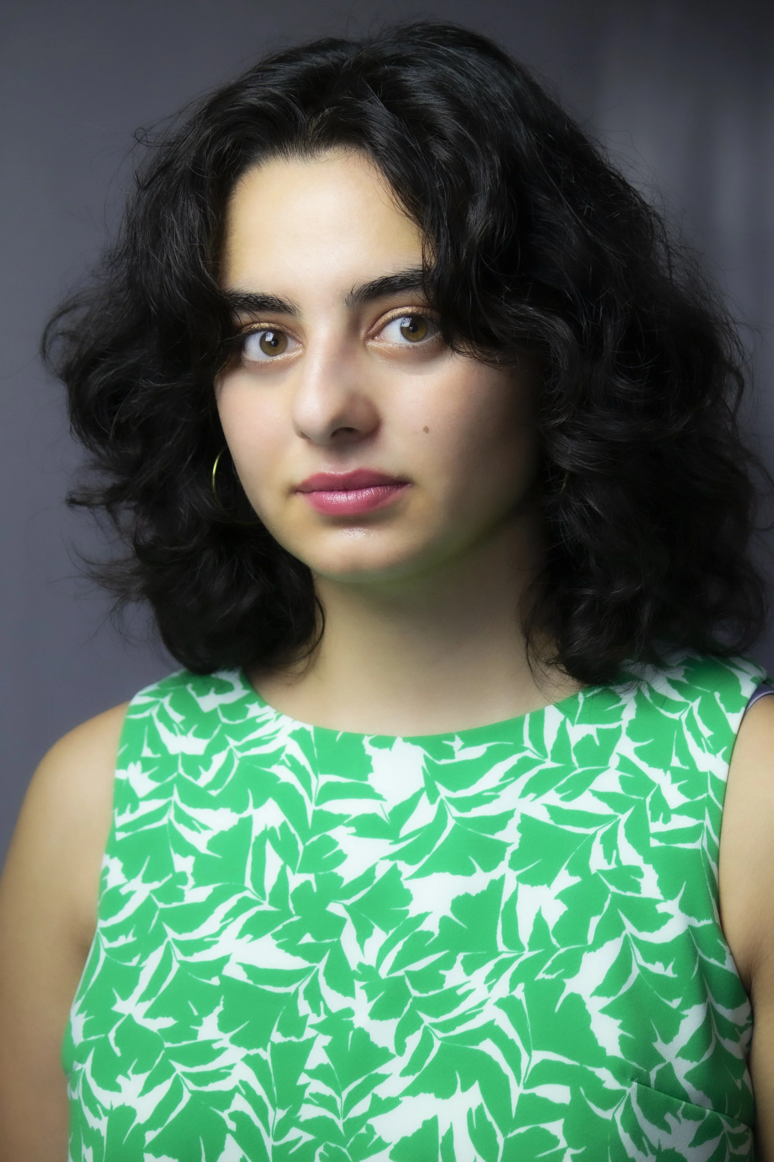 Portrait of a young woman with dark curly hair wearing a green and white patterned sleeveless top, against a dark gray background.