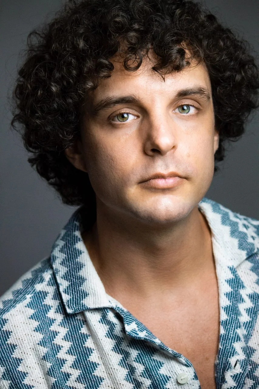 A man with curly dark hair and light eyes, wearing a blue and white patterned shirt, looking directly at the camera with a neutral expression.