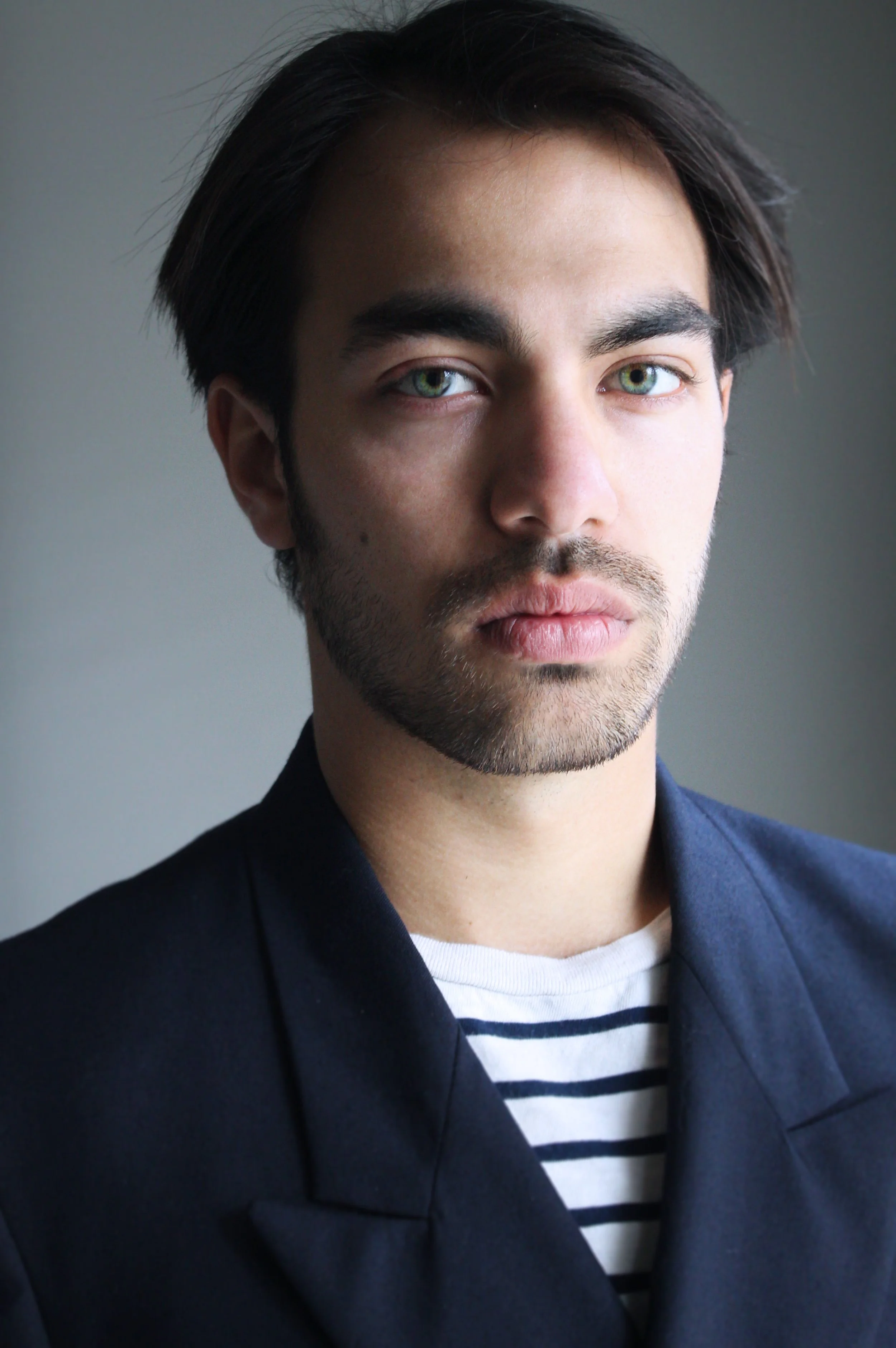 A young man with dark hair, light skin, and blue eyes, wearing a dark blazer over a striped white and black shirt, looking at the camera with a neutral expression.