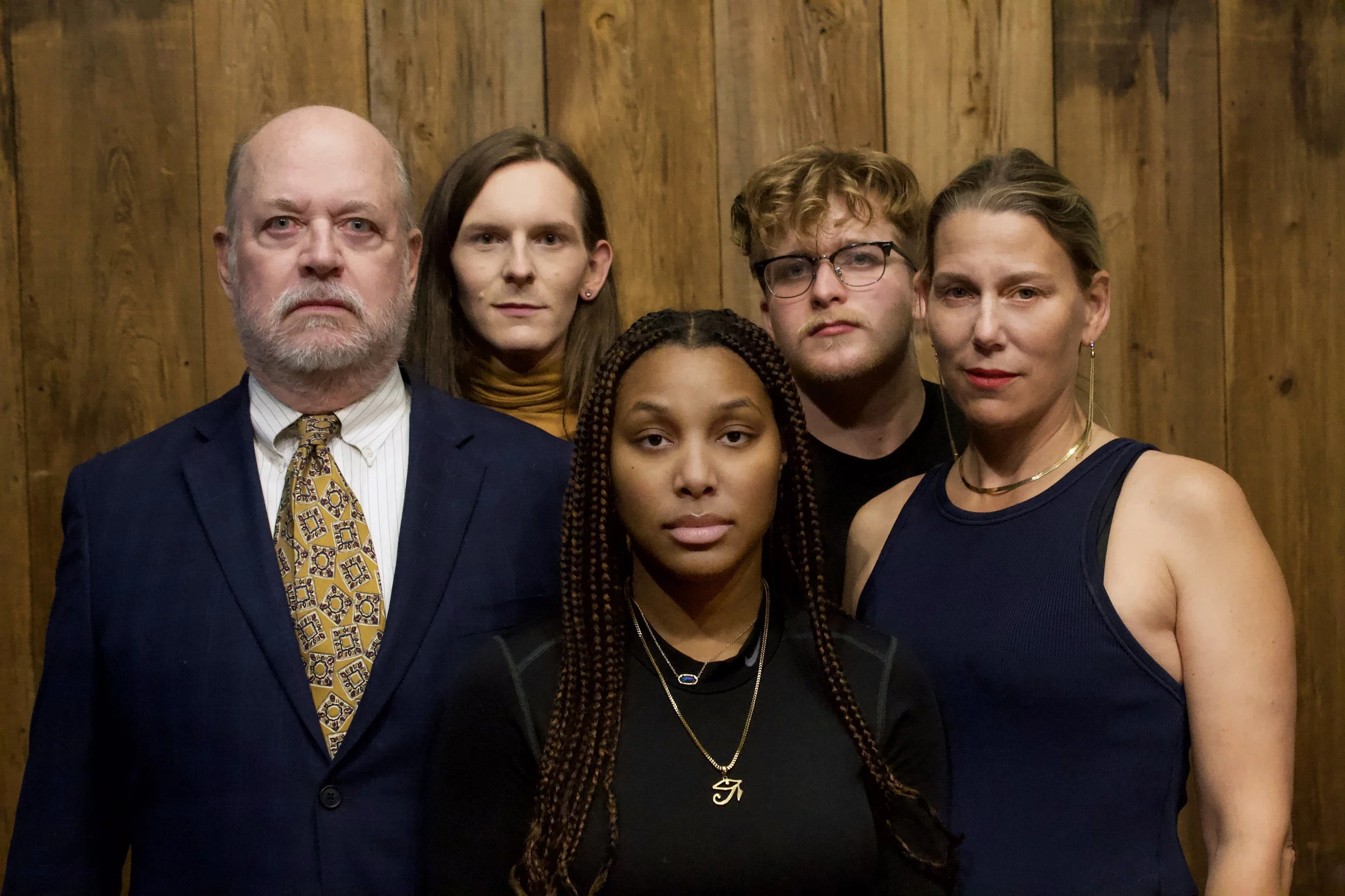A group of six diverse people standing against a wooden wall, looking directly at the camera.