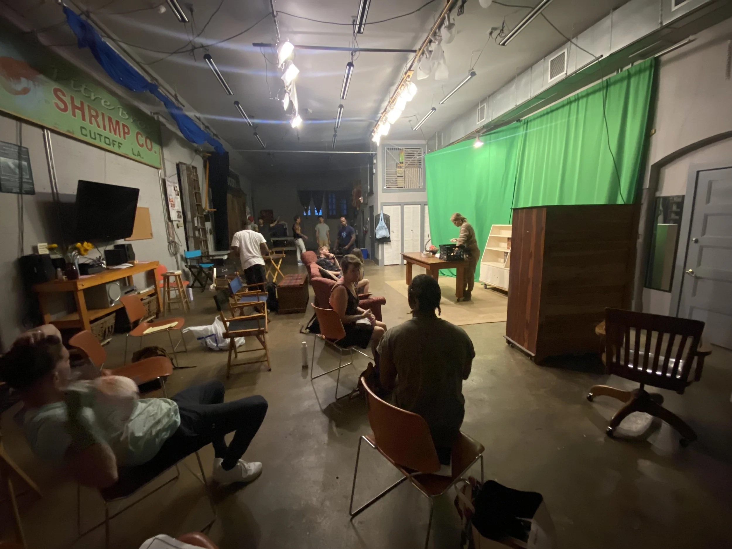 People waiting in a room with a green screen on the wall, some seated, some standing, with a woman working at a desk.