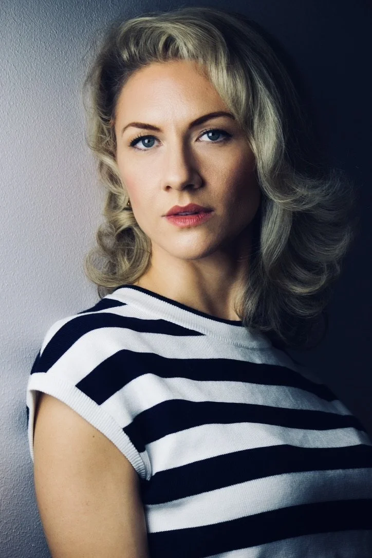 A woman with shoulder-length blonde hair and blue eyes wearing a black and white striped shirt, posing against a background split into light and dark.