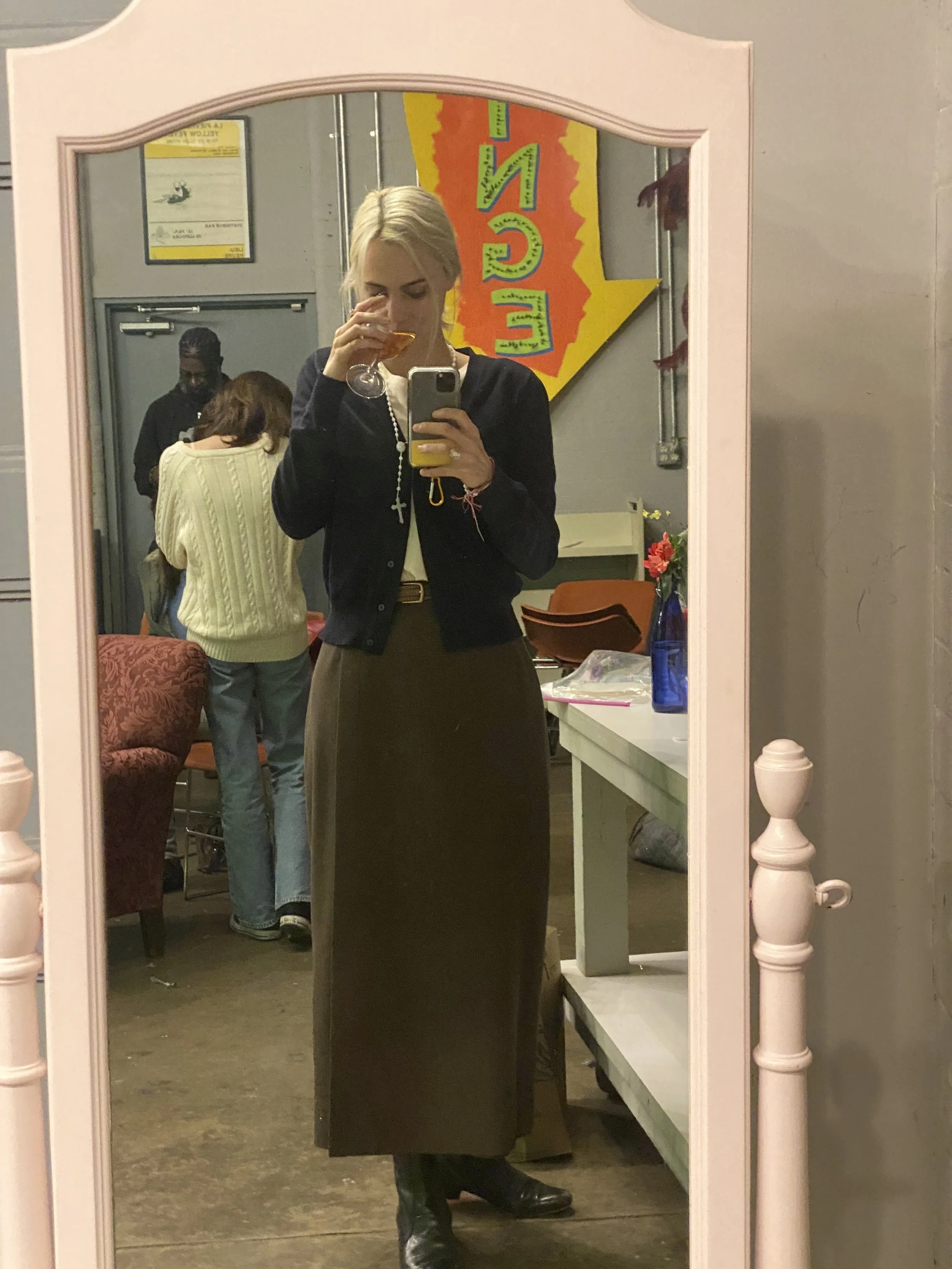 A woman taking a selfie in a mirror, wearing a long dark skirt, black jacket, and boots, holding a wine glass, with a cross necklace and a phone with a yellow case. There are two people in the background, one in a yellow sweater and another in black,