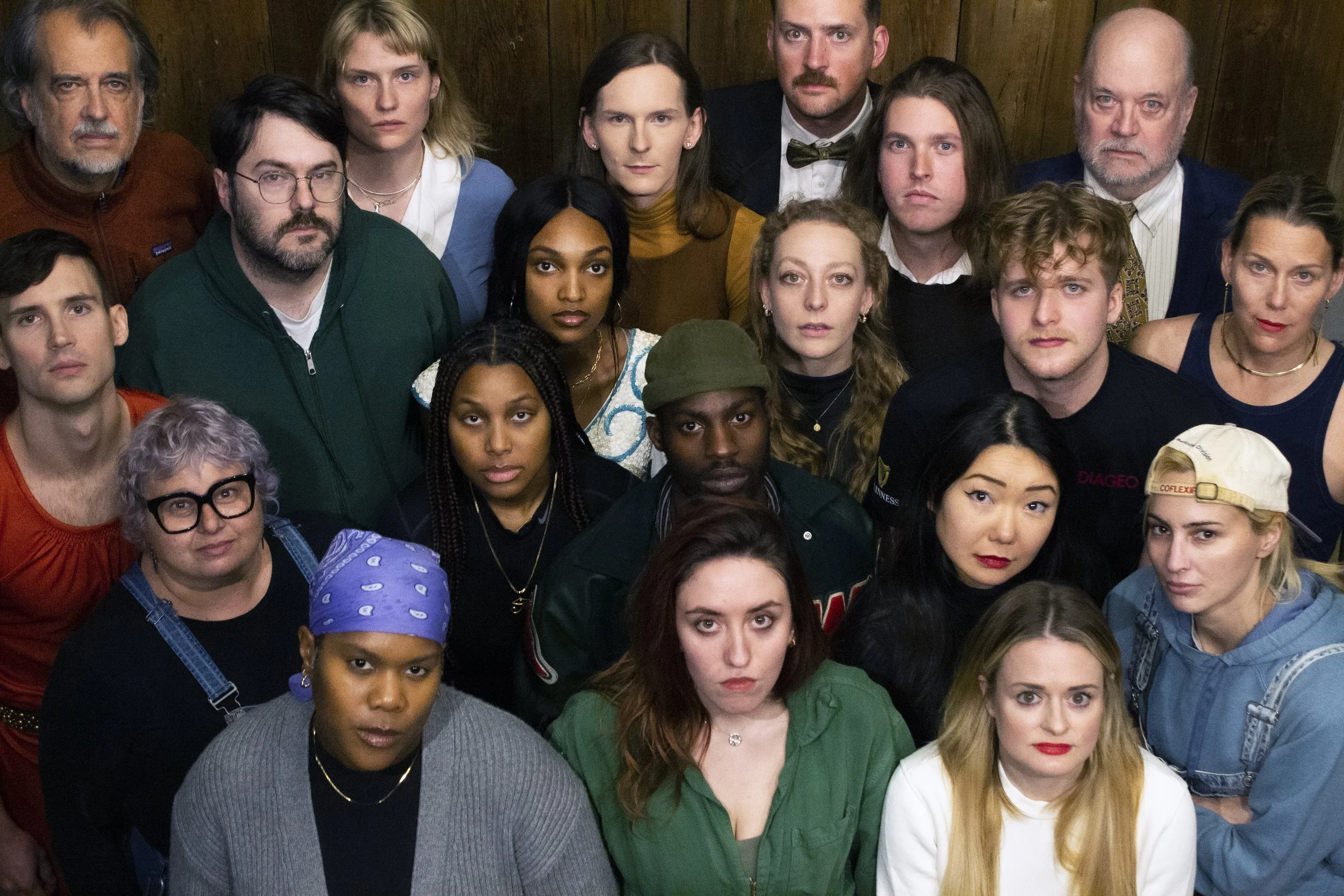 Group of diverse people posing together in a close-up photo. The group includes men and women of different ages and ethnicities, standing indoors with a wooden background.