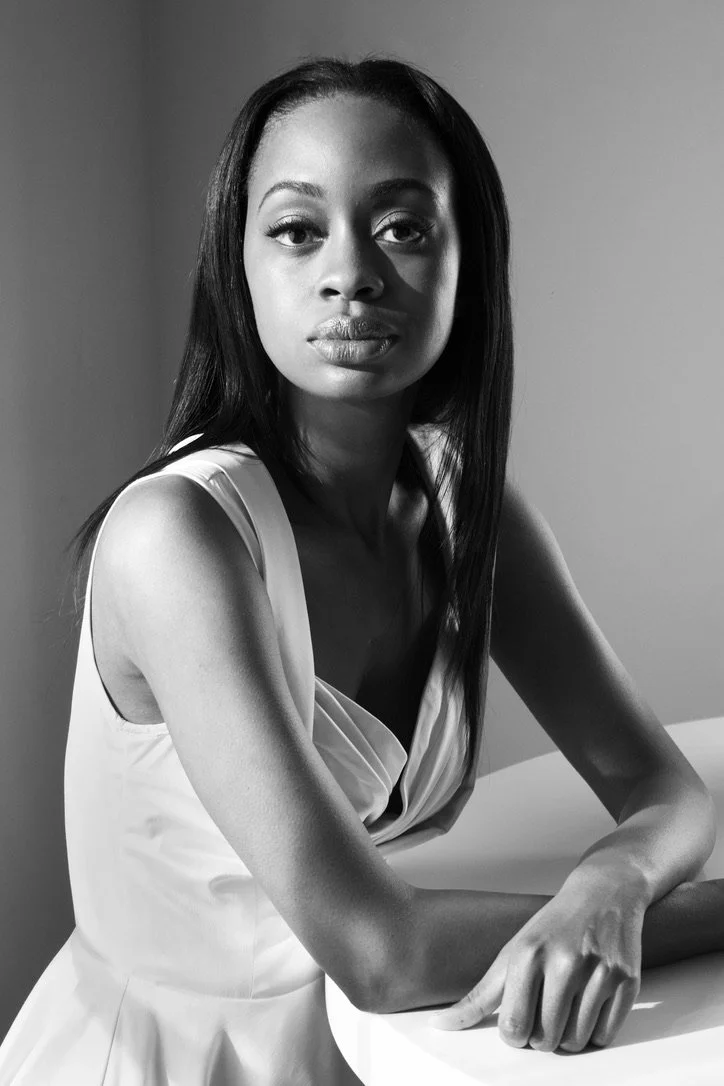 Black and white portrait of a woman with long straight hair, wearing a sleeveless dress, seated with her arms resting on a surface and looking directly at the camera.