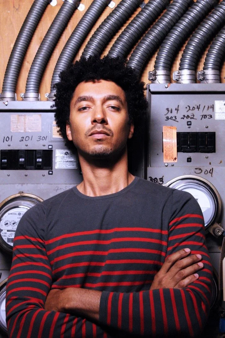 A man standing with his arms crossed in front of electrical meters and wiring in a utility room or basement.