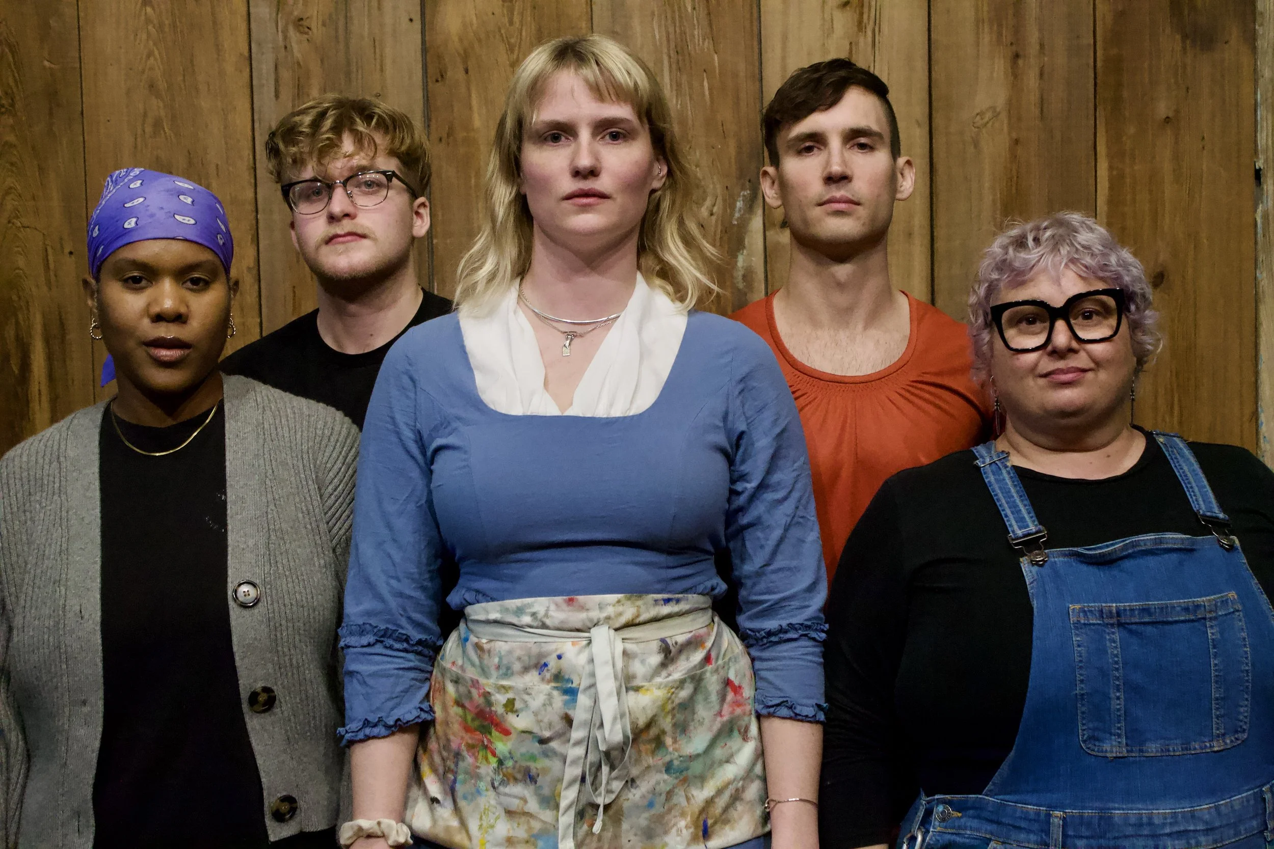Group of five diverse people standing against a wooden wall, looking serious at the camera.