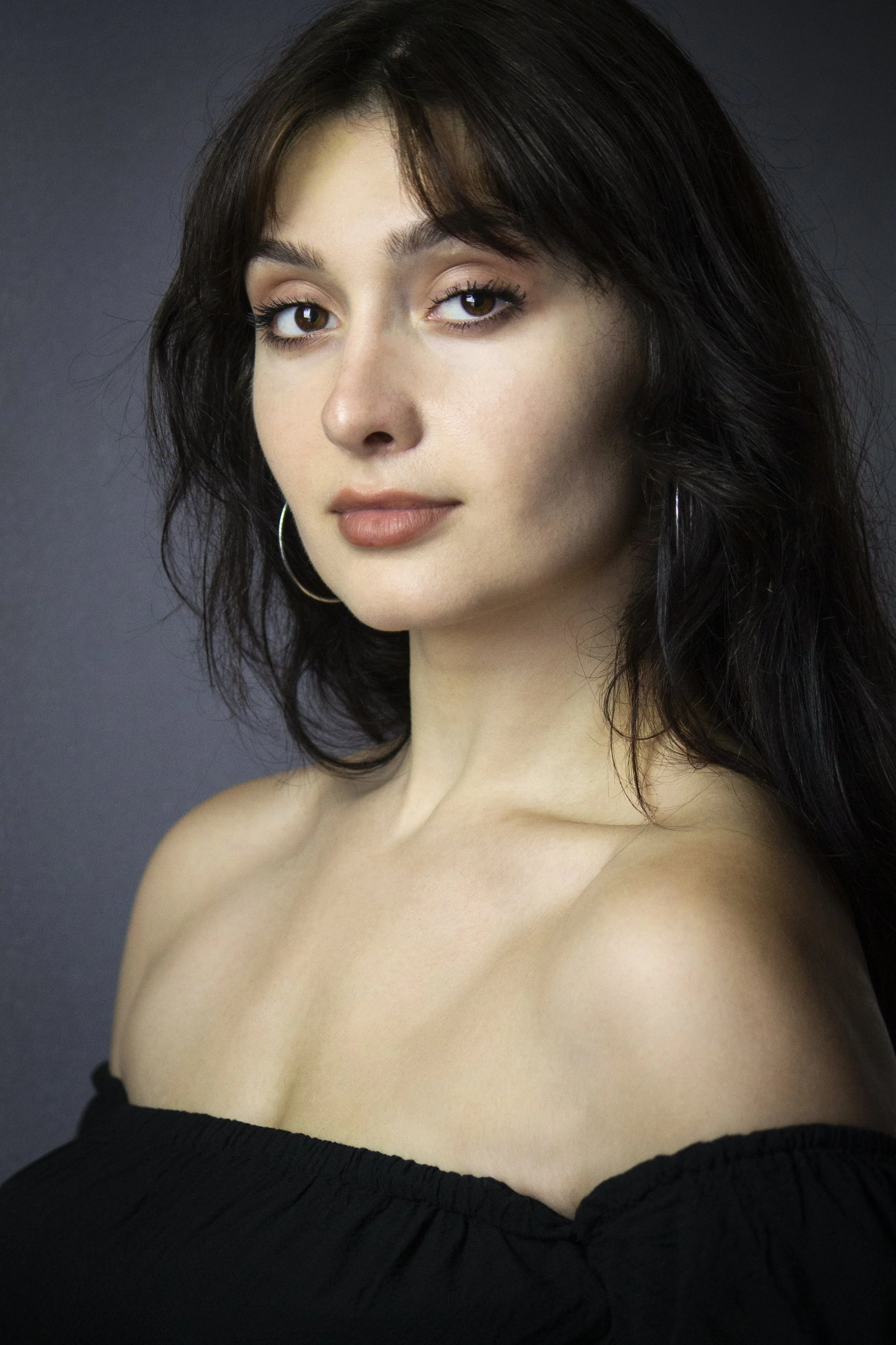 A young woman with fair skin, dark wavy hair, and brown eyes wearing hoop earrings and a black off-the-shoulder top, looking at the camera against a gray background.