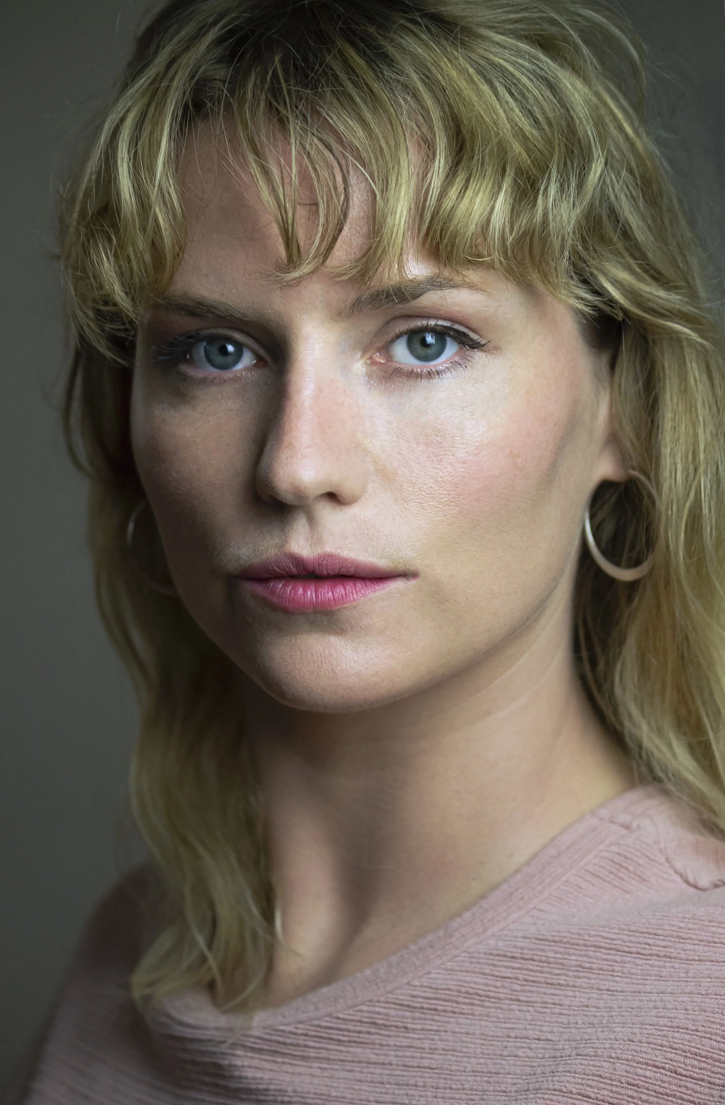 Close-up portrait of a woman with blonde, wavy hair, blue eyes, wearing a pink top and hoop earrings.