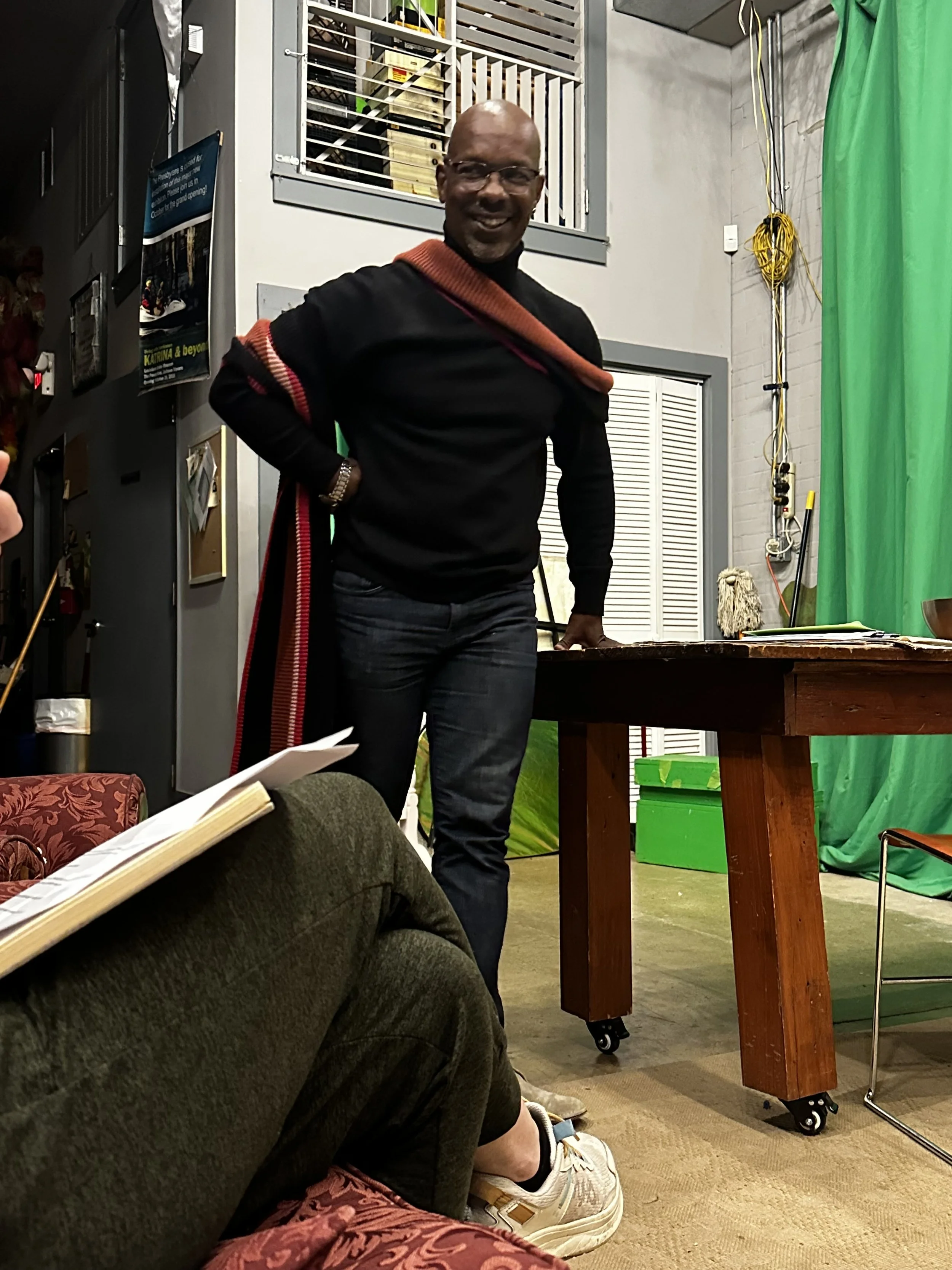 A man in a black sweater with a red scarf stands smiling in front of a green screen and a table, in a room with various equipment and decorations.