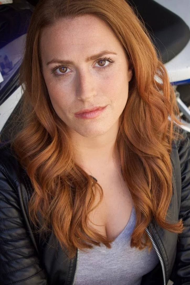 A woman with long, wavy red hair and fair skin is looking directly at the camera. She is wearing a black leather jacket over a gray shirt.
