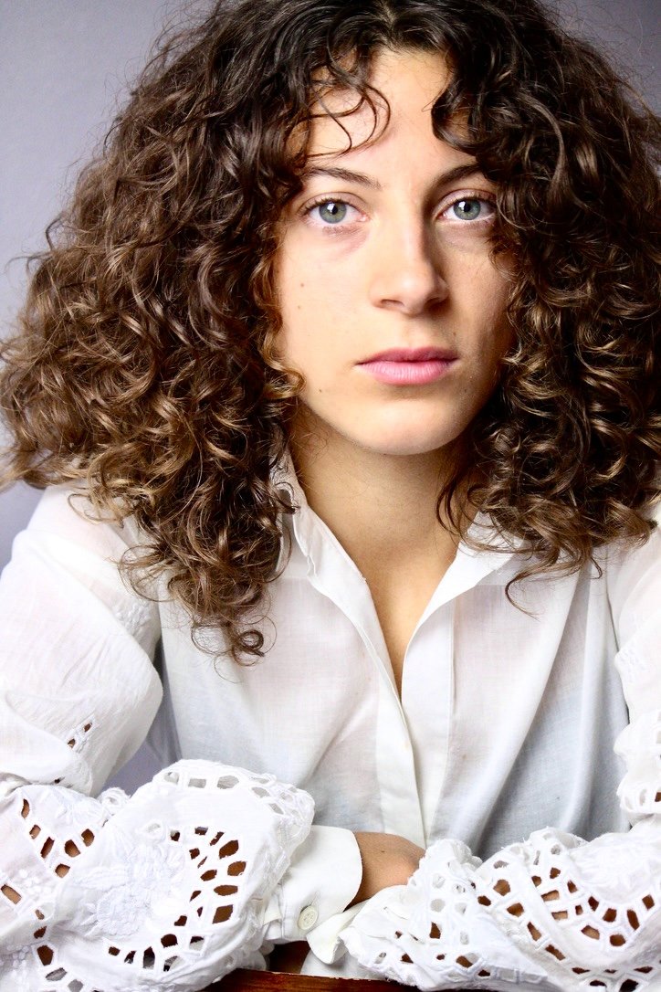 Close-up portrait of a young woman with curly brown hair and blue eyes wearing a white shirt with lace details on the sleeves.
