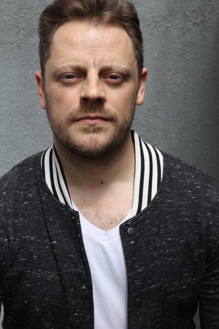 A man with short brown hair, light skin, and a beard, wearing a black jacket with white and black striped collar over a white t-shirt, standing against a gray wall.