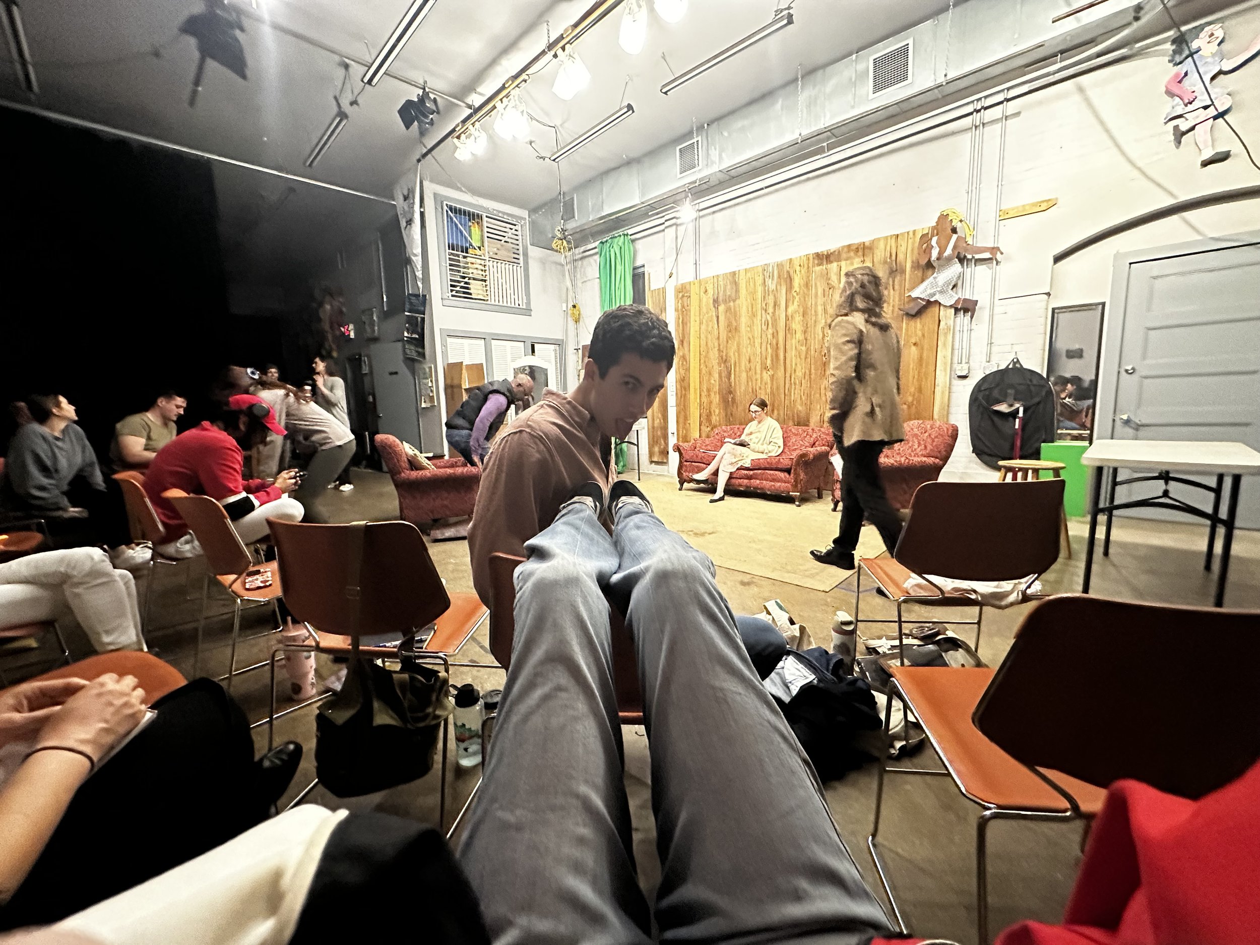 People seated and standing in a theater or rehearsal space with a stage area, wooden backdrop, and various props and decorations on the walls.