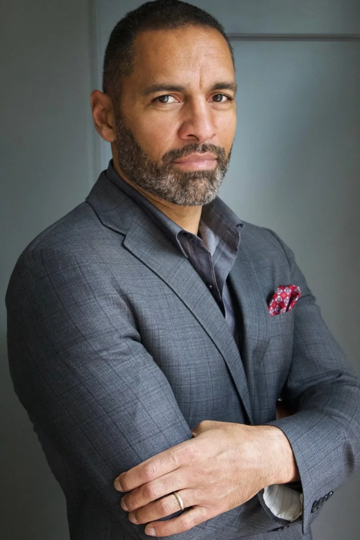 A man in a gray suit with a red pocket square standing with arms crossed, looking directly at the camera.