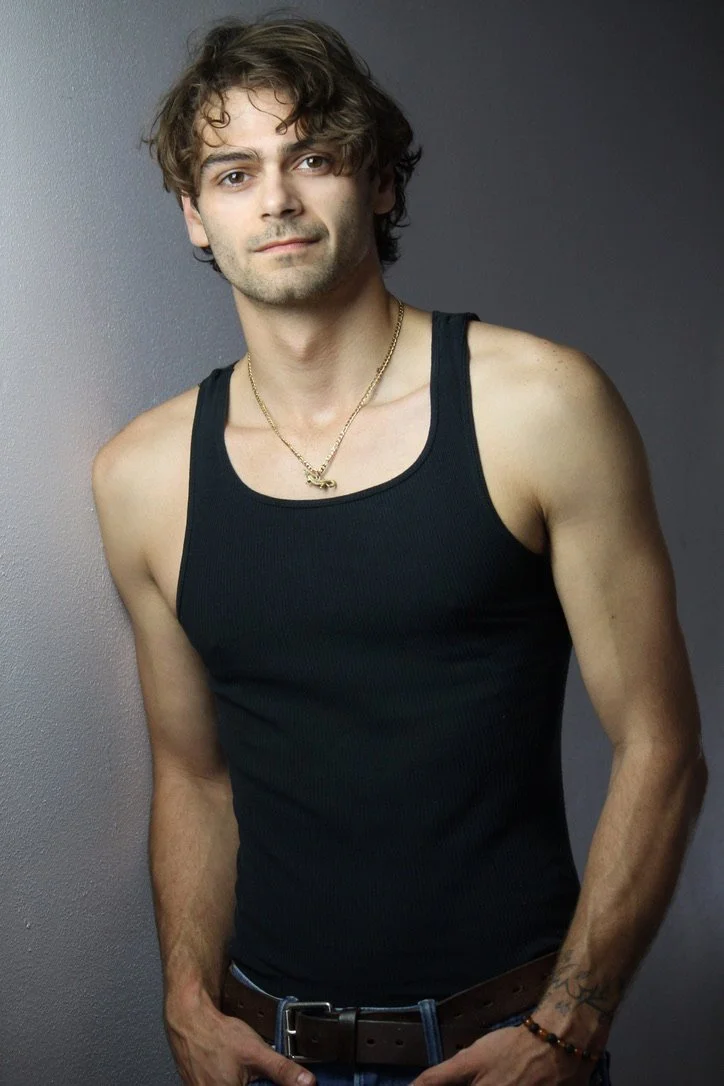 Young man with wavy brown hair, wearing a black tank top, a gold necklace, and a bracelet, standing against a gray background.