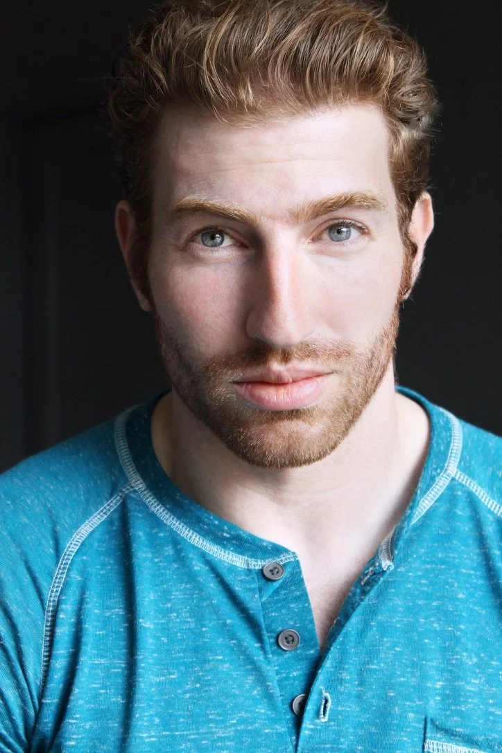 A young man with reddish-blonde hair and blue eyes looking directly at the camera. He has a light beard and is wearing a blue, long-sleeve shirt with buttons.