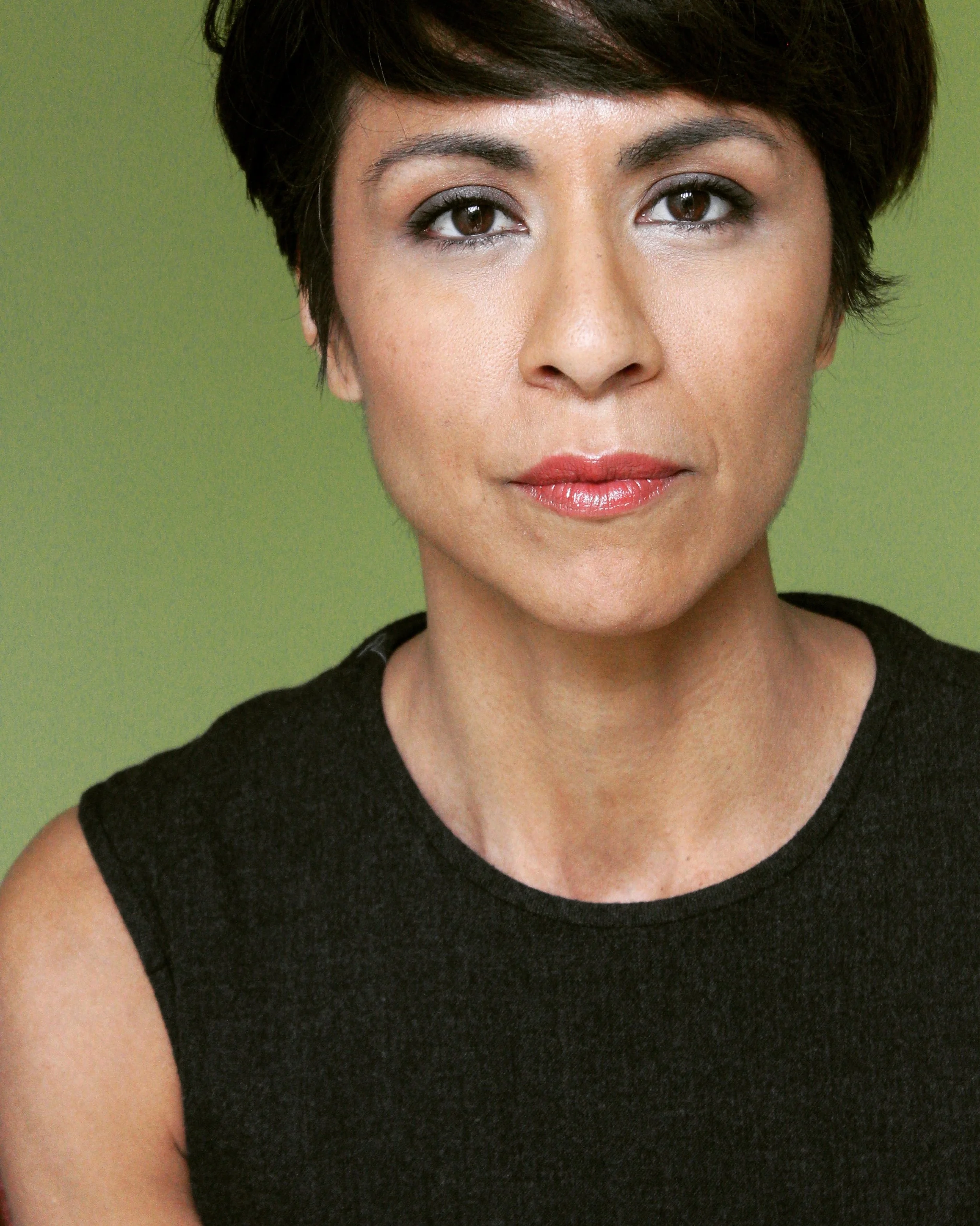 Close-up of a woman with short dark hair, wearing a black sleeveless top, against a green background.