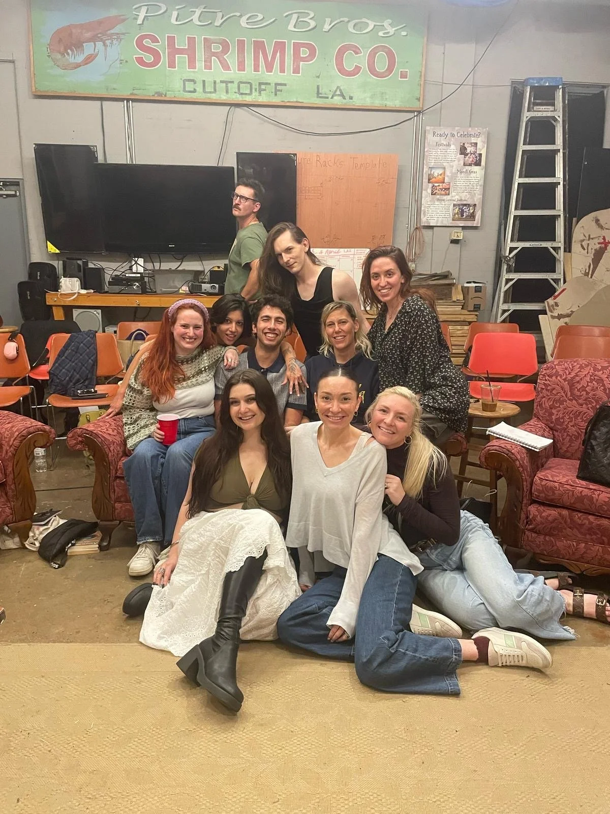 Group of nine people gathered indoors, sitting and kneeling on the floor and couches, smiling for a photo in front of a wall with a large piano and a sign that reads 'Pitre Bros. SHRIMP CO. CUT OFF LA'.