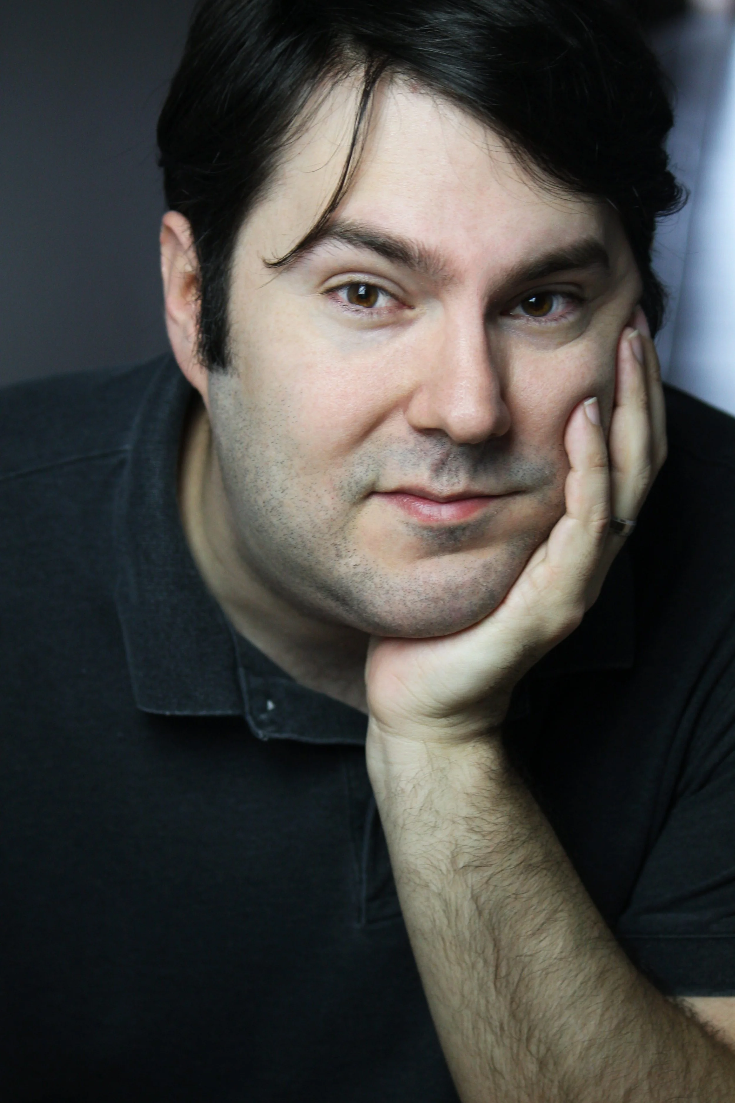 Close-up portrait of a man with dark hair, resting his face on his hand, wearing a black collared shirt.
