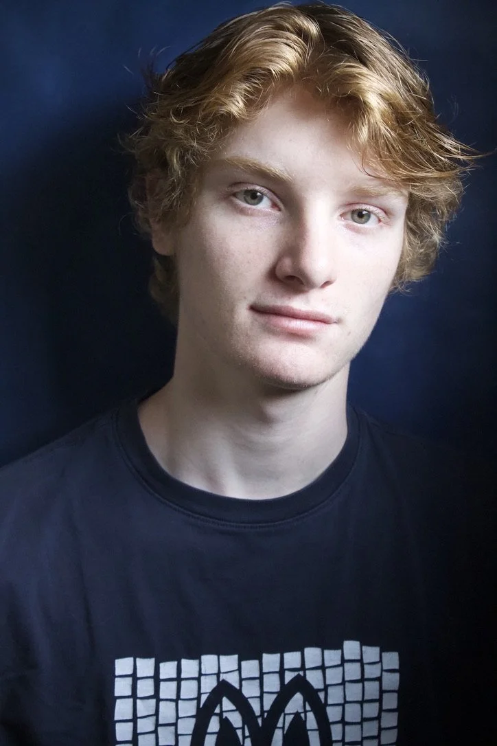 A young man with red hair and blue eyes posing against a dark blue background, wearing a black T-shirt with a white graphic design.