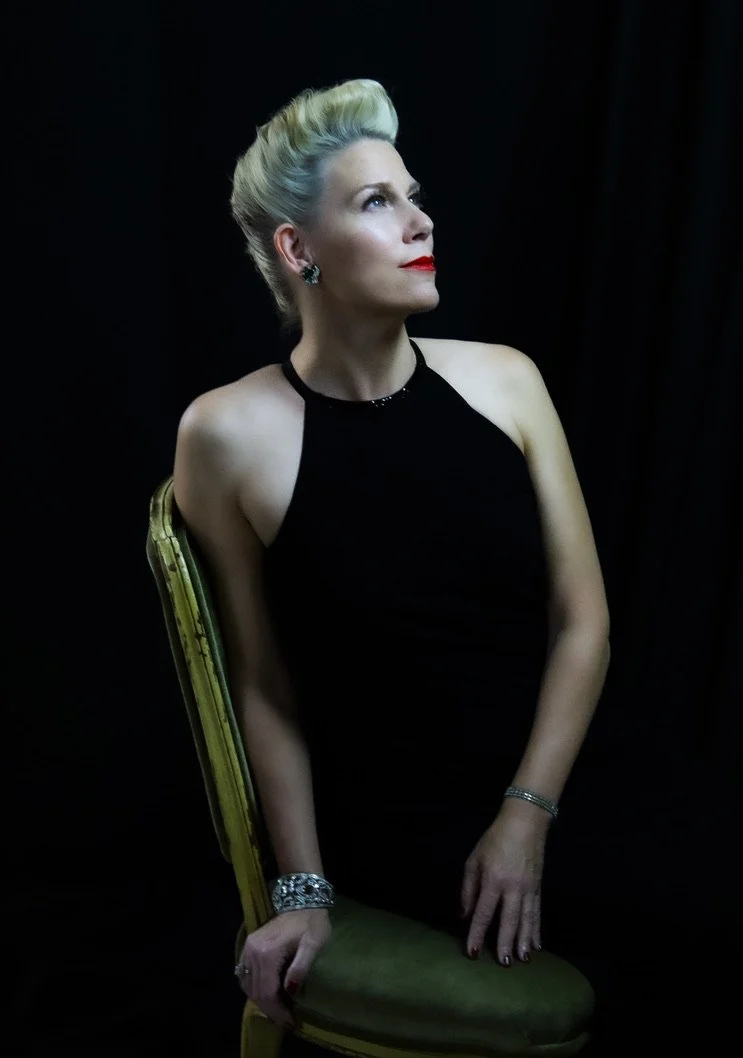 A woman dressed in a black halter dress with platinum blonde hair styled up, sitting on a green velvet chair with a dark background, looking to her right with a confident expression, wearing red lipstick, earrings, and bracelets.