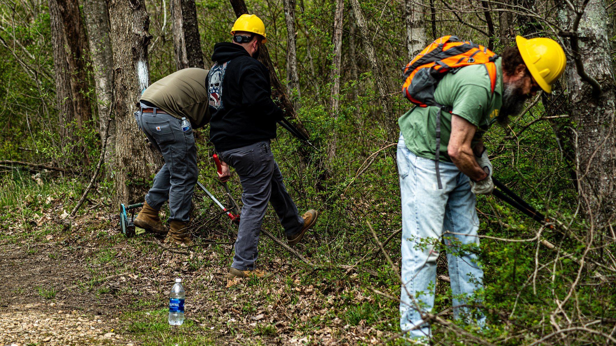 Three men wearing yellow helmets and outdoor clothes working together in a wooded area, using metal detectors to search for something underground.