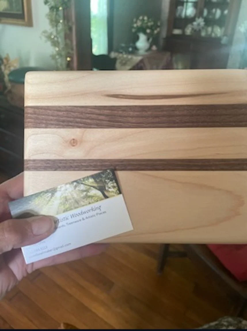 A hand holding a business card and a wooden cutting board with light and dark wood stripes in a room with wooden floors, plants, and antique furniture.