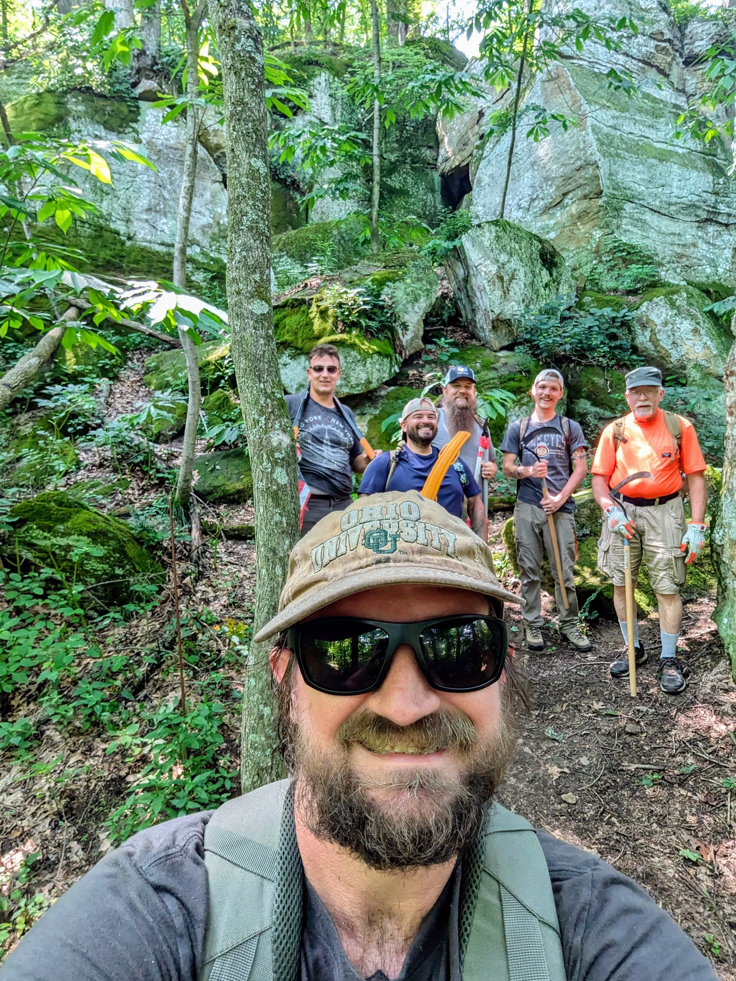 A group of five men in outdoor clothing and backpacks standing and smiling in a forested area with large rocks and green foliage, with the person in the foreground taking a selfie. The photo is taken in a densely wooded area with moss-covered rocks.