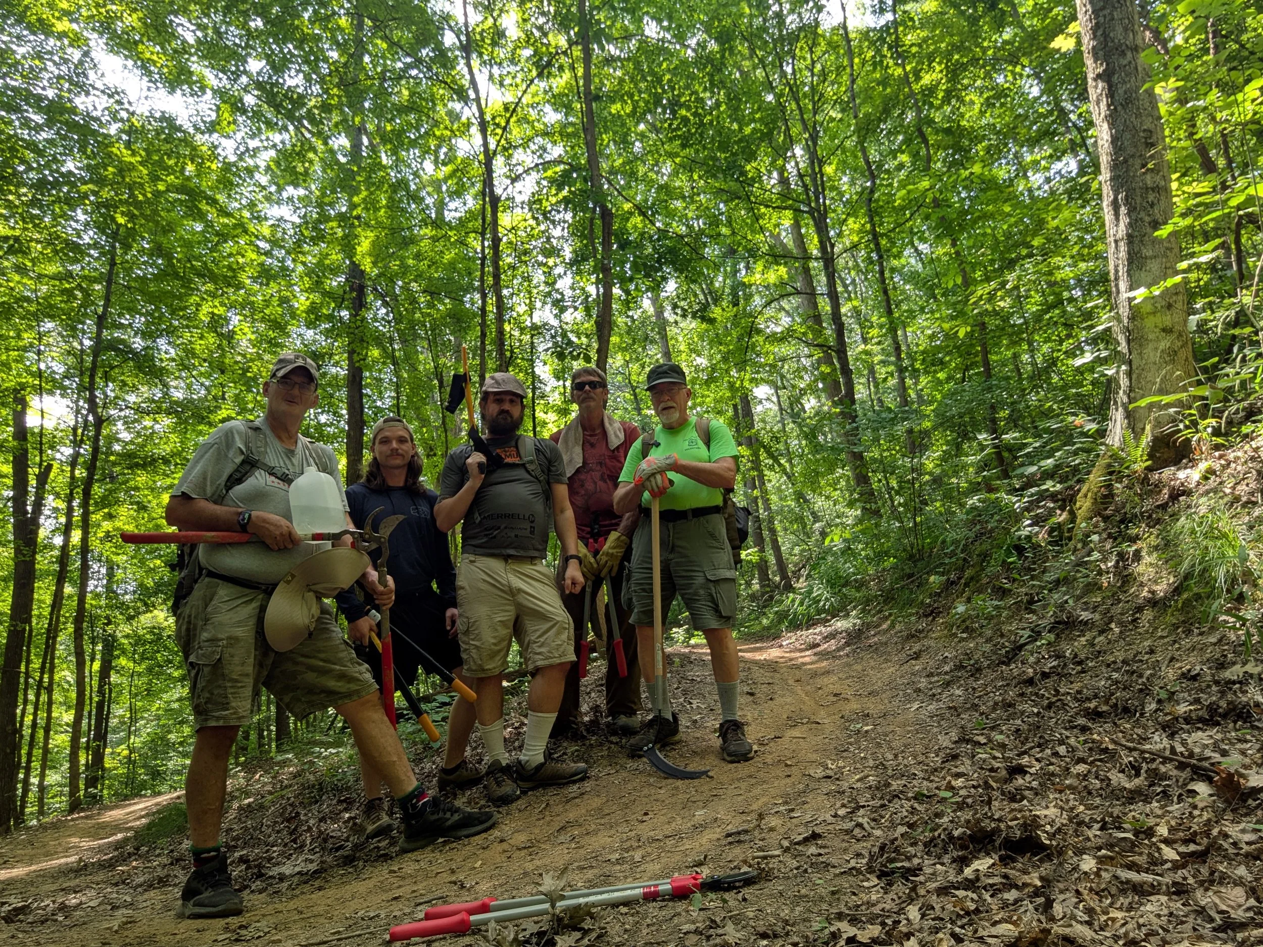 Group of five hikers standing on a dirt trail in a dense green forest, some holding tools and wearing backpacks.