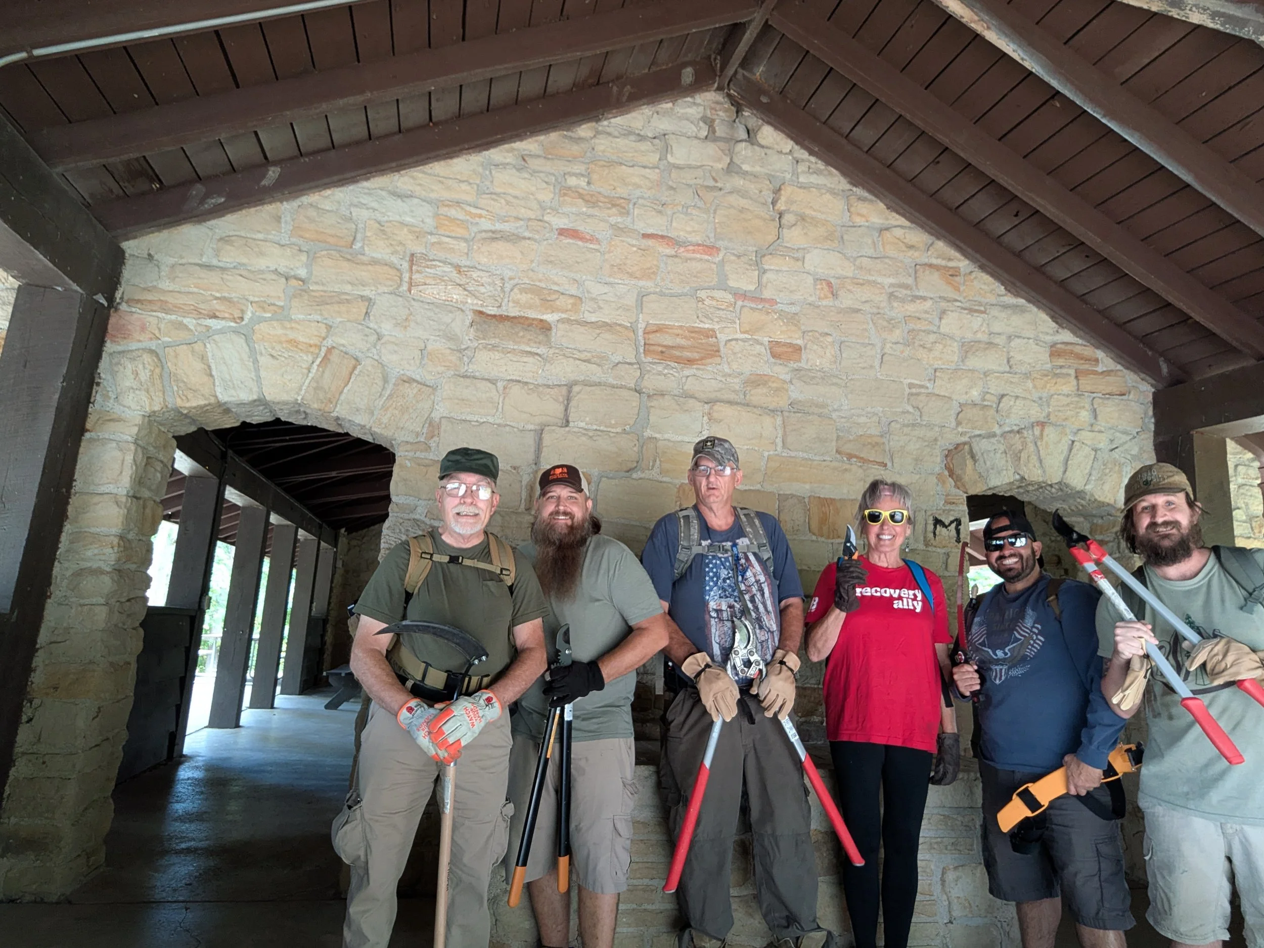 Group of six people smiling and holding tools, standing in front of a stone wall and wooden ceiling.