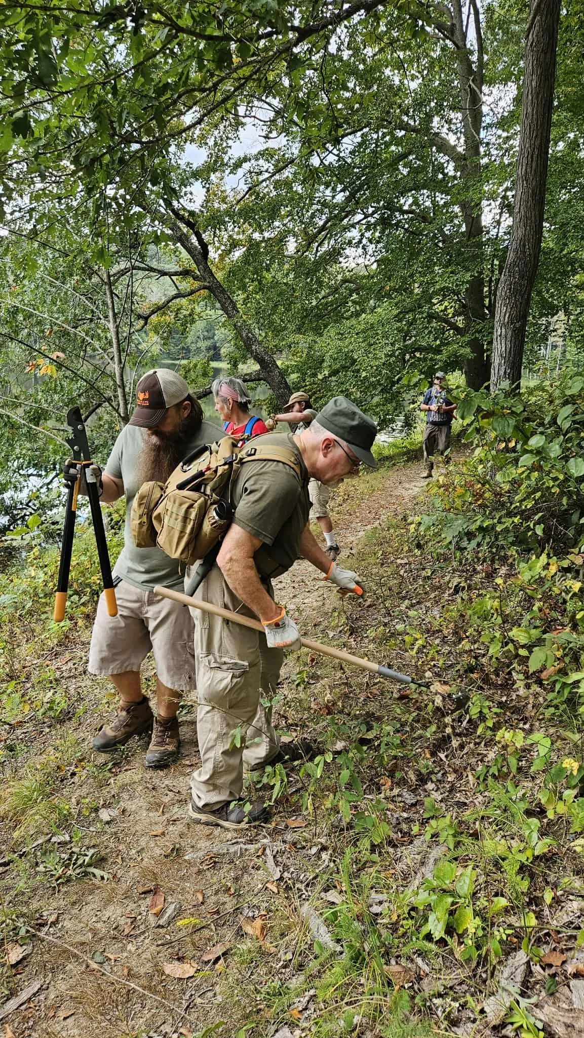 A group of people on a nature trail, using tools for plant or soil clearing, surrounded by trees and greenery.