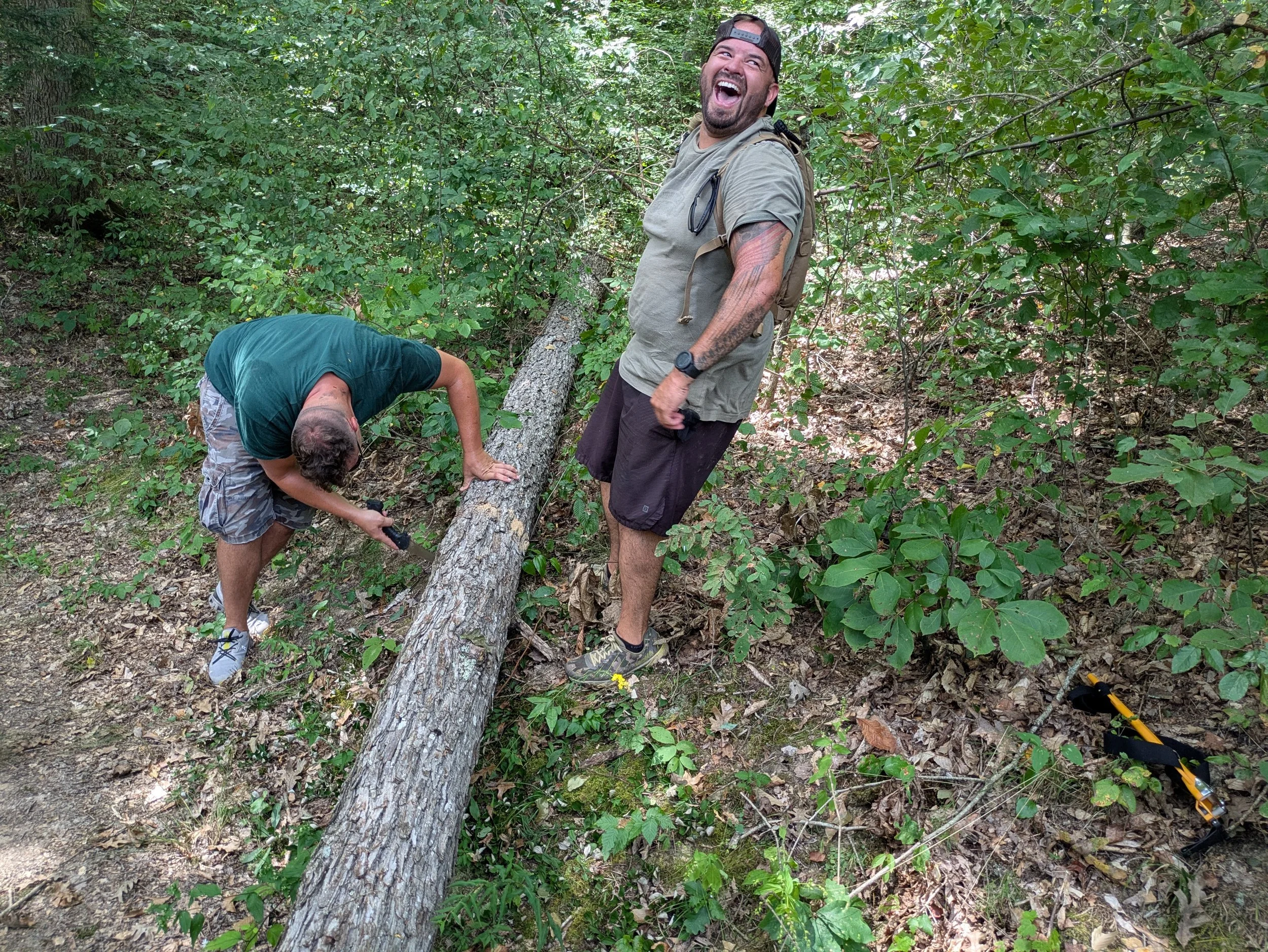 Two men in a wooded area, one bending down and using a saw on a fallen tree, the other standing and laughing, holding a phone, with lush green foliage all around.