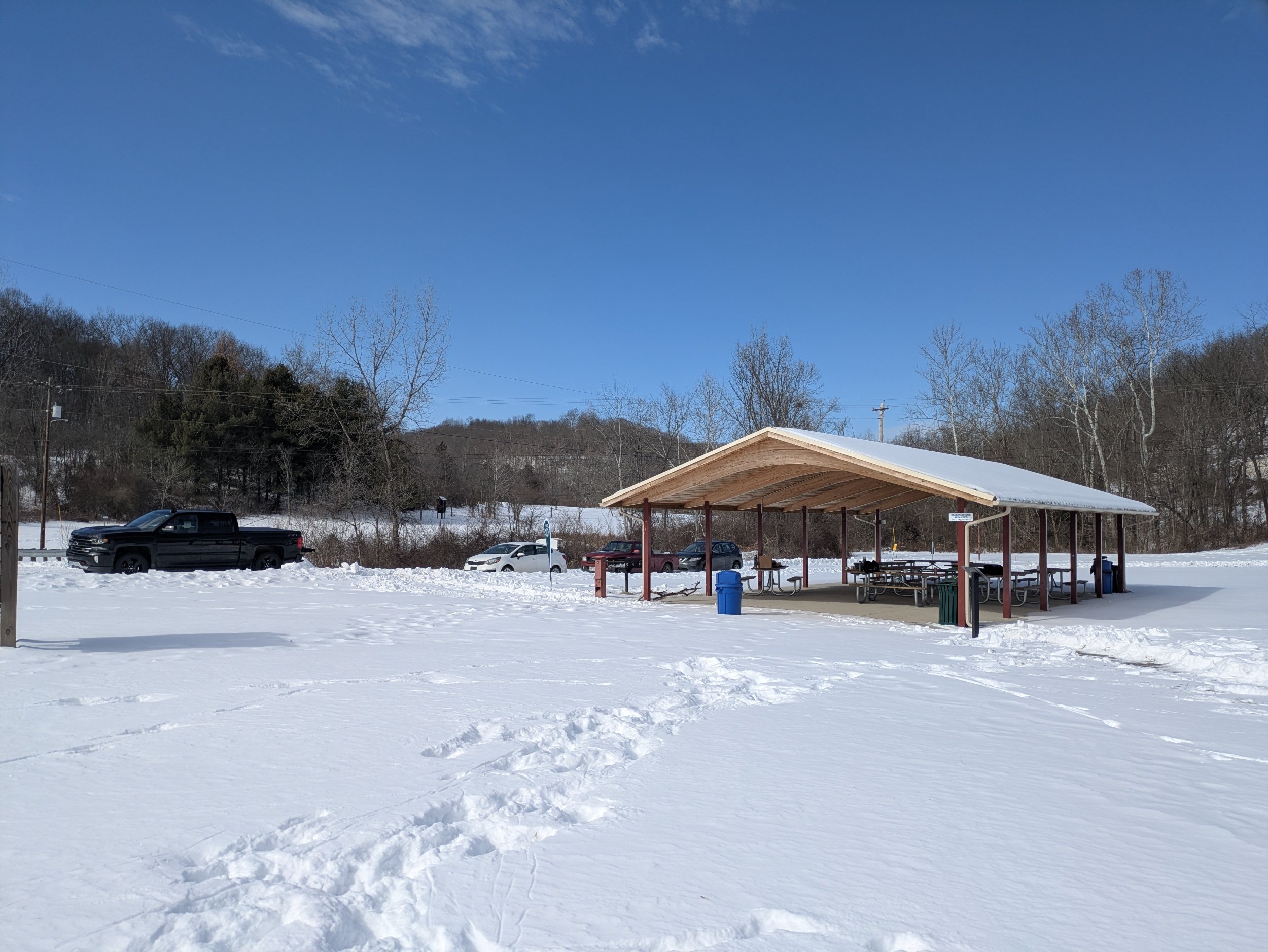 Snow-covered parking lot with a wooden shelter containing picnic tables, surrounded by leafless trees on a clear winter day.