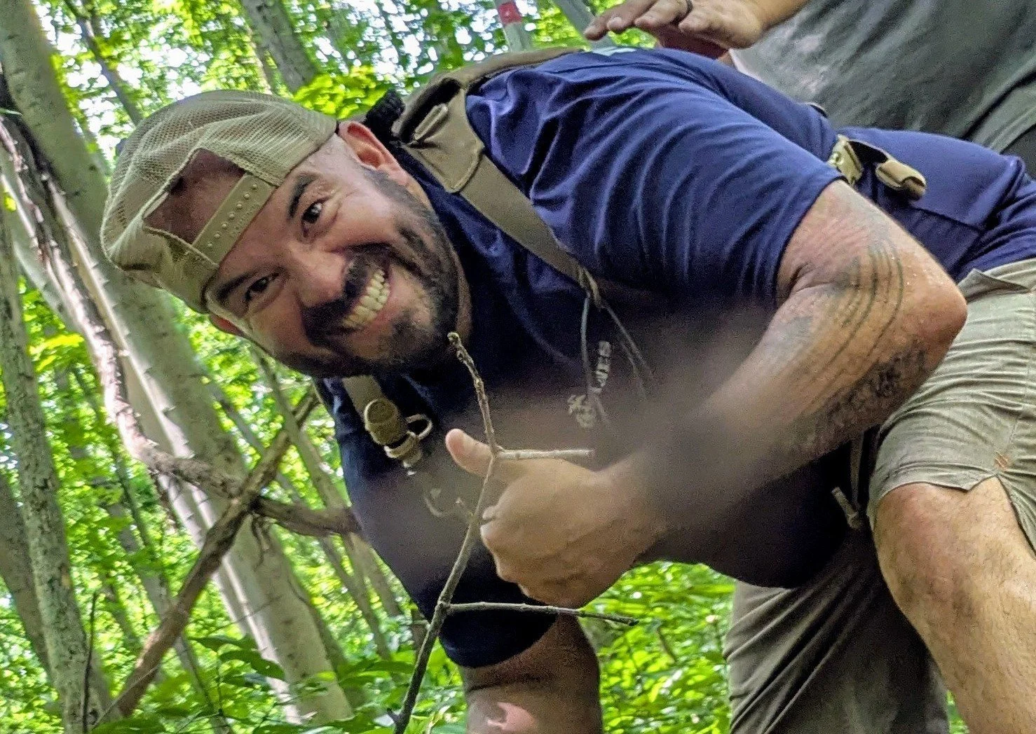 A man smiling and giving a thumbs-up while outdoors in a forest, crouching near trees with green leaves, wearing a tan cap, navy blue shirt, and khaki shorts, with a backpack strap over his shoulder.