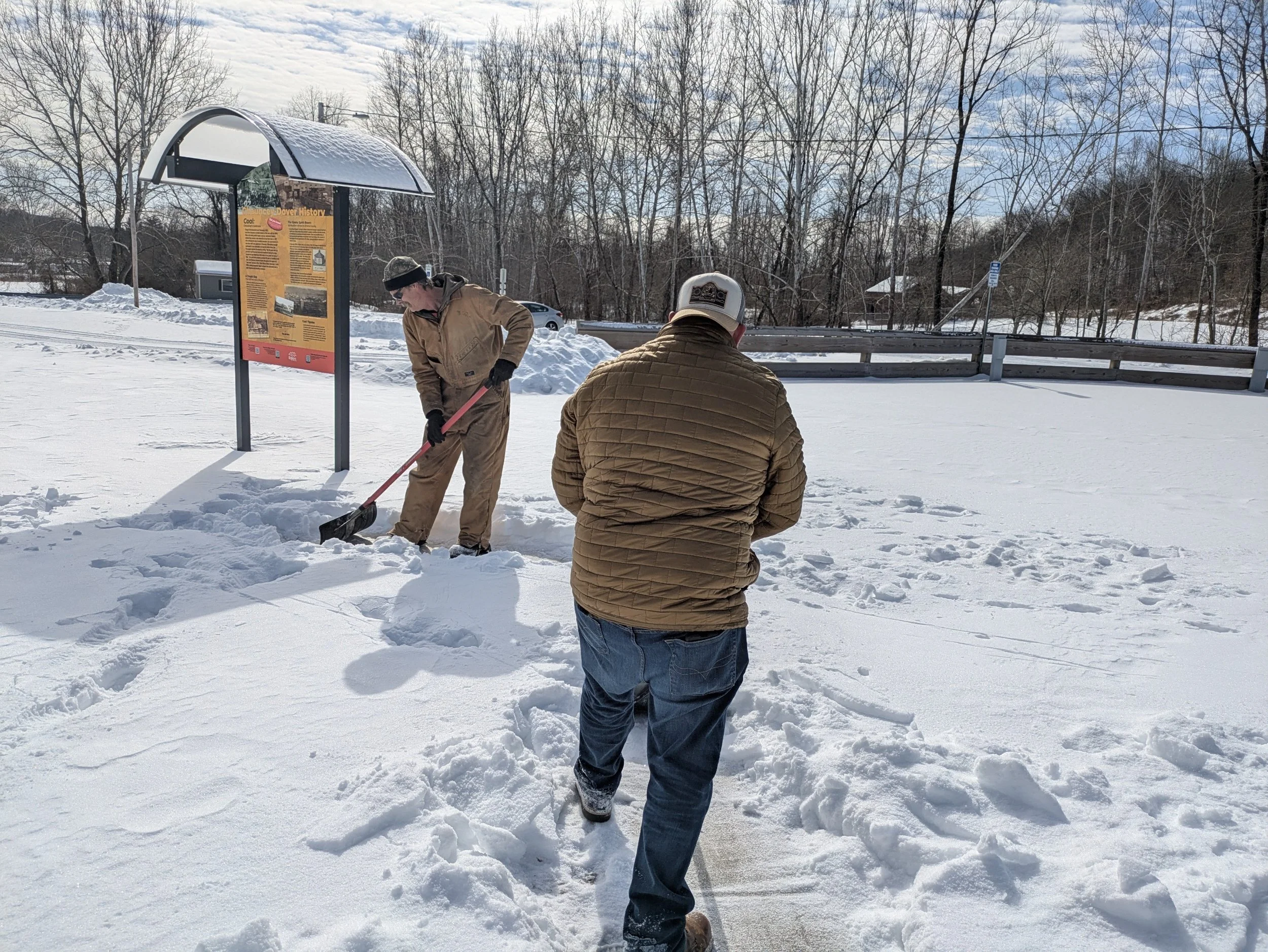 Two men are standing on snow-covered ground, one shoveling snow while the other looks at a smartphone. In the background, there is a wooden fence, a utility pole, and a bulletin board with a cover, under a partly cloudy sky.