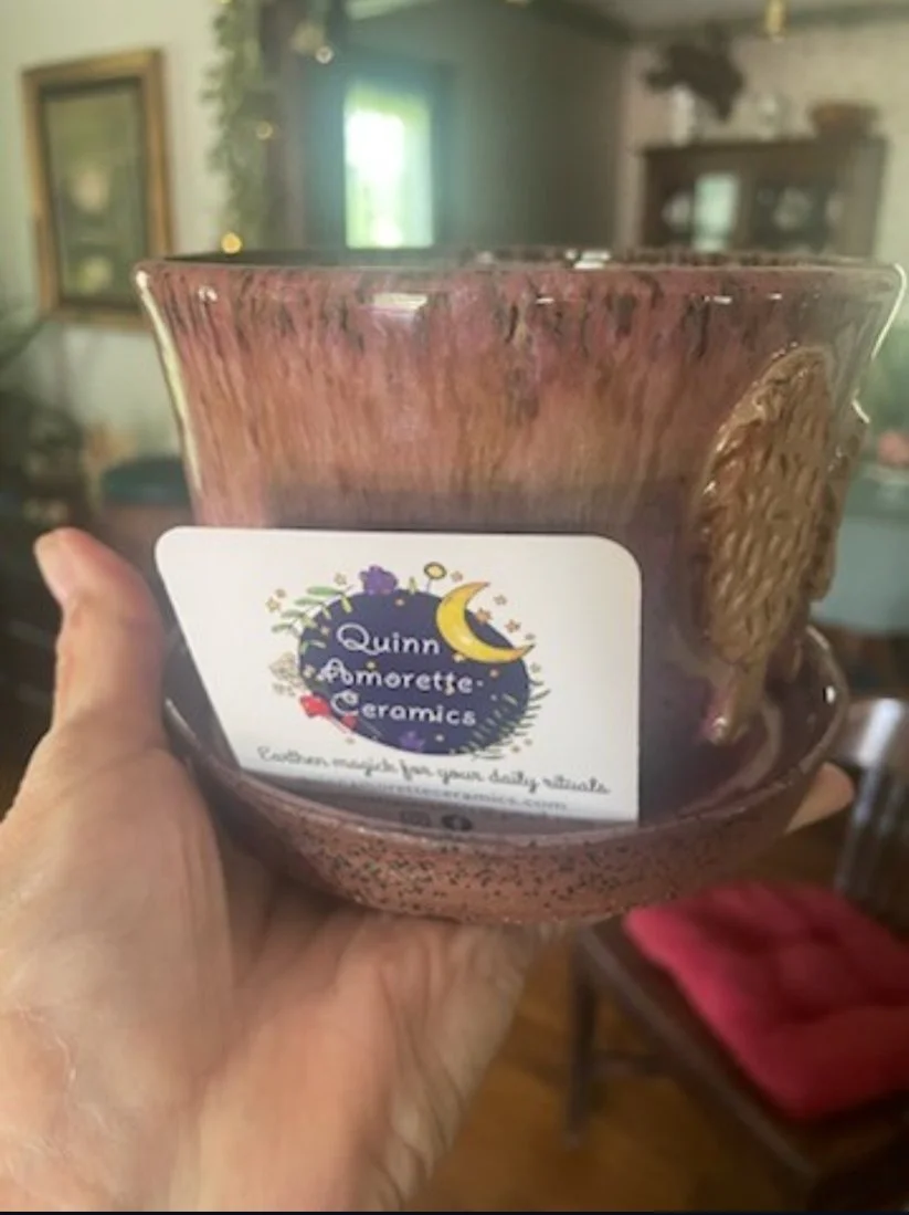 A hand holding a ceramic bowl with a colorful label that reads, "Quinn Amorette Ceramics" with a moon and star design. The bowl has a textured, reddish-brown glaze and is set in a cozy room with wooden furniture and dark walls.