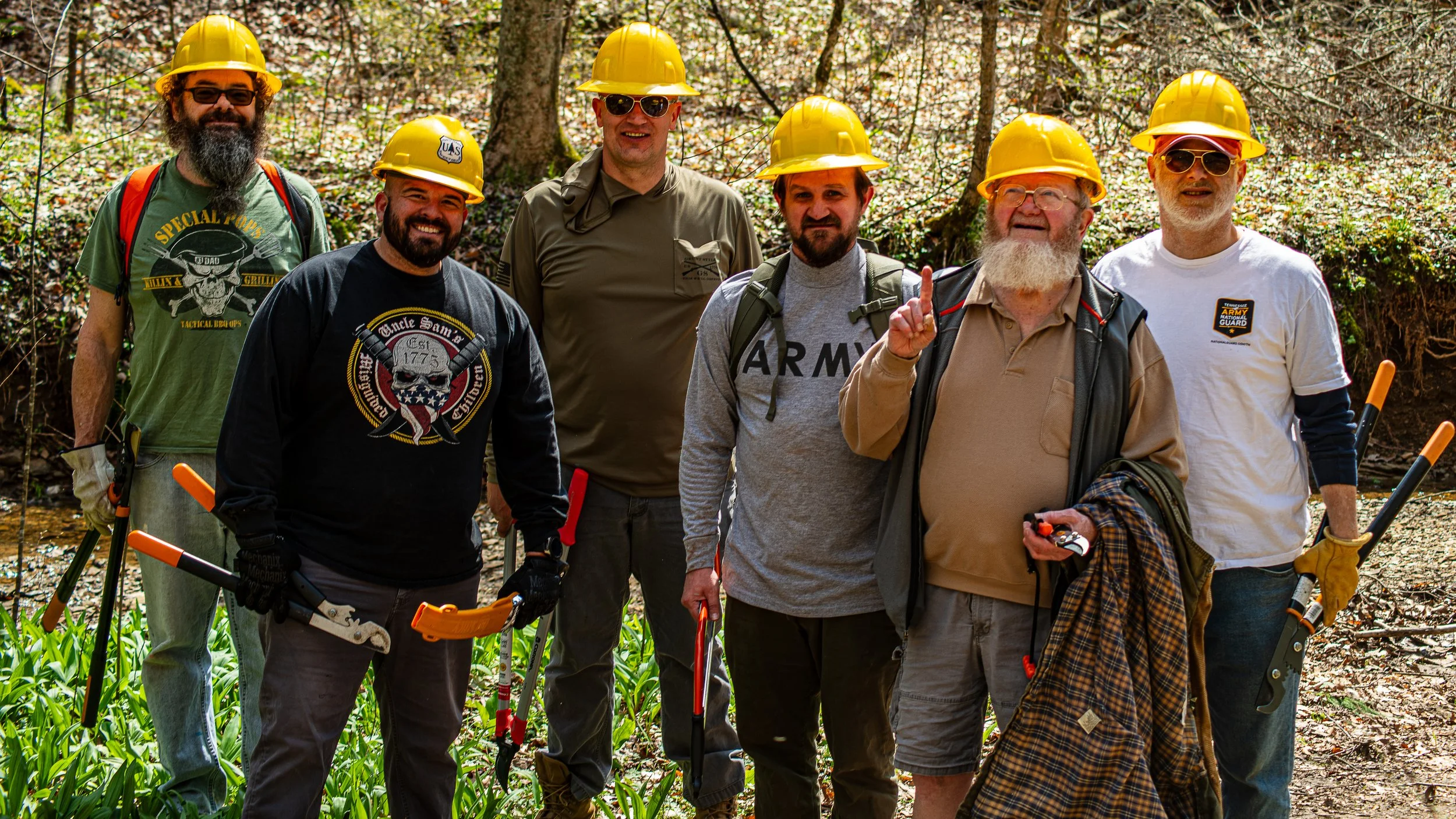 Group of seven men wearing yellow safety helmets standing outdoors in a wooded area, holding tools for land clearing or tree cutting.