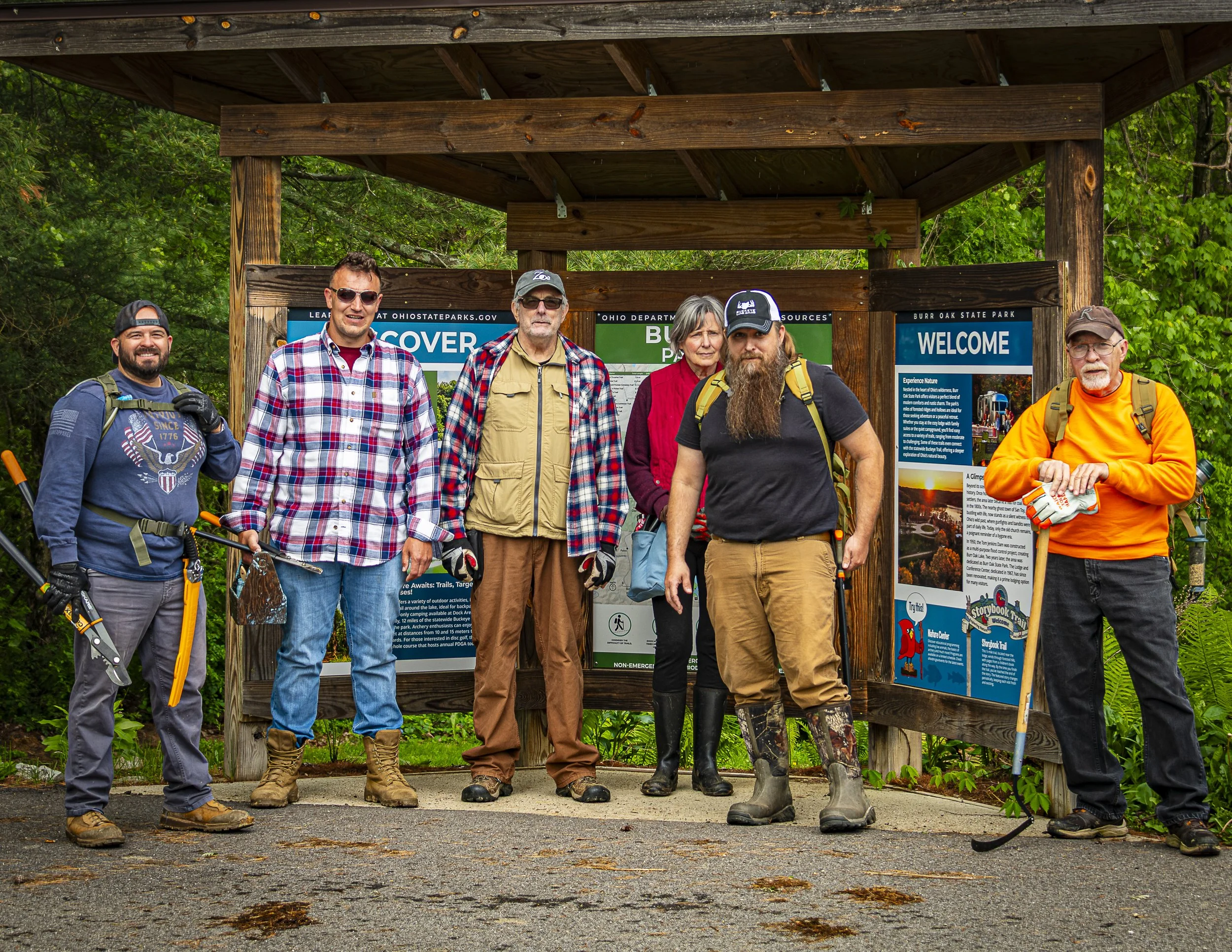 Group of six hikers standing in front of an information board at a park, surrounded by trees and greenery.