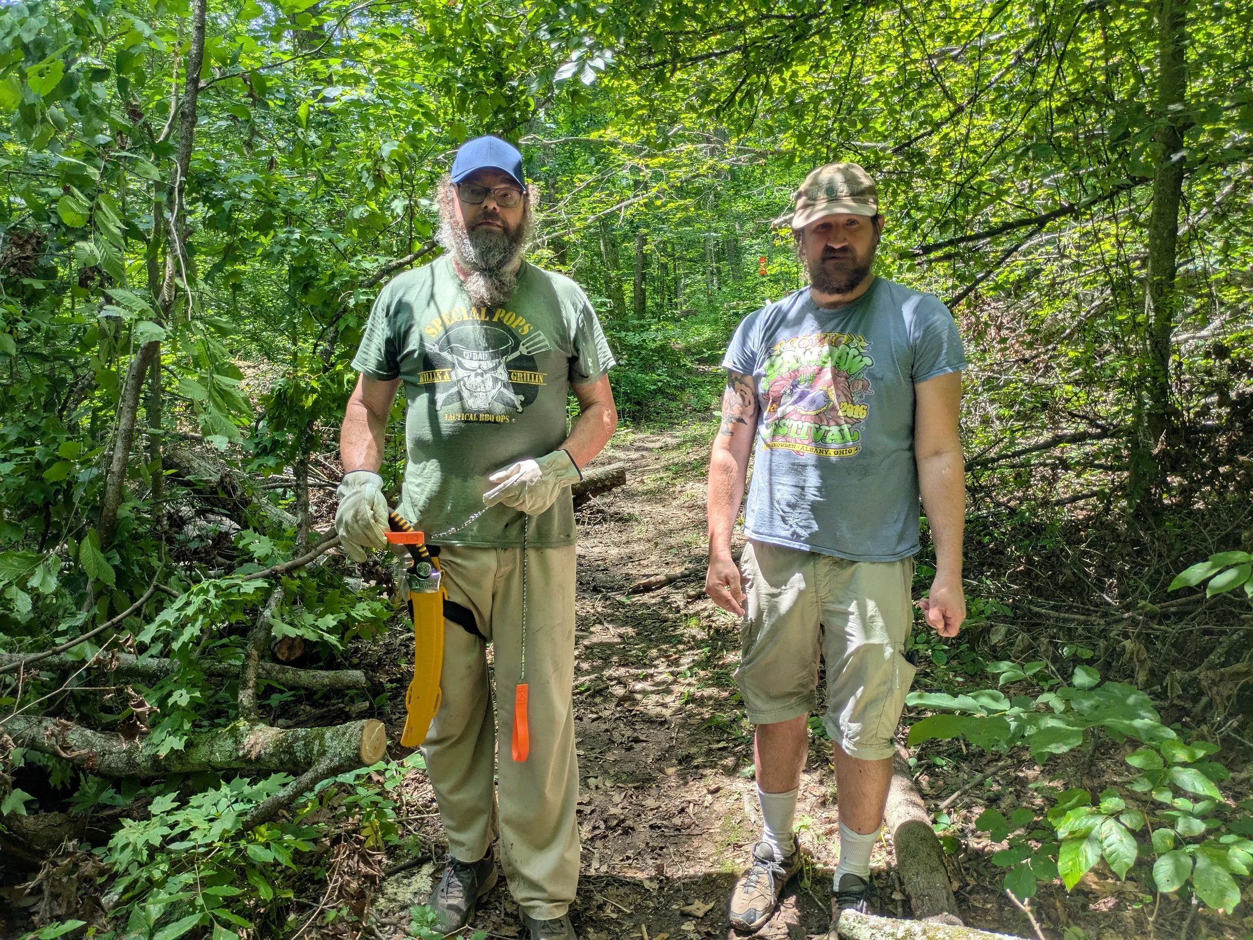 Two men standing in a wooded area during daylight. The man on the left has a beard, sunglasses, a blue cap, and is holding a saw. The man on the right is wearing a camo cap, a graphic t-shirt, and shorts. They appear to be engaged in outdoor work or 