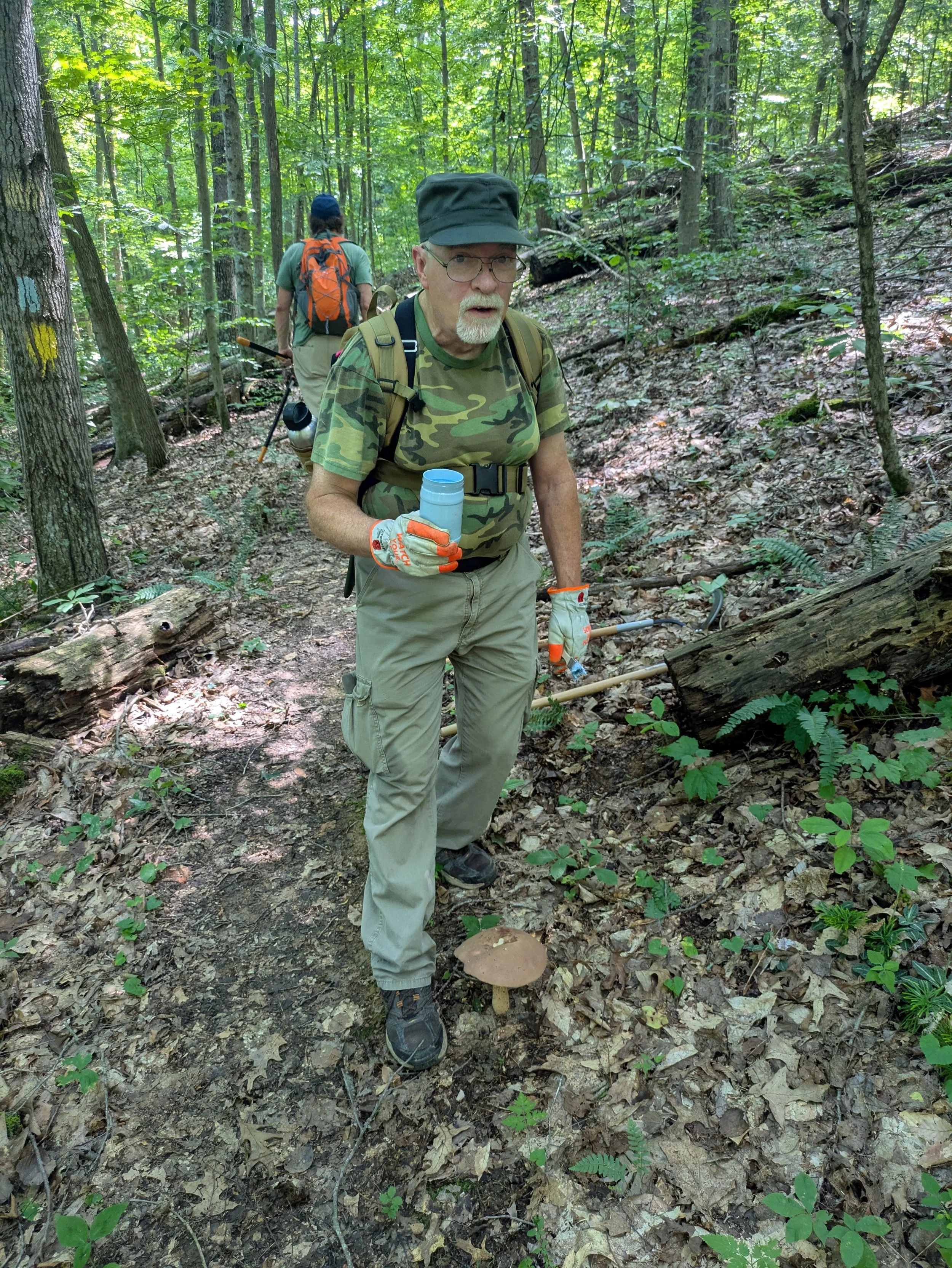 Two men on a trail in a dense, green forest; the man in front wears a camouflage t-shirt, khaki pants, gloves, glasses, and a green hat, holding a cup; the man in back has an orange backpack, a green shirt, and khaki shorts, carrying a walking stick.