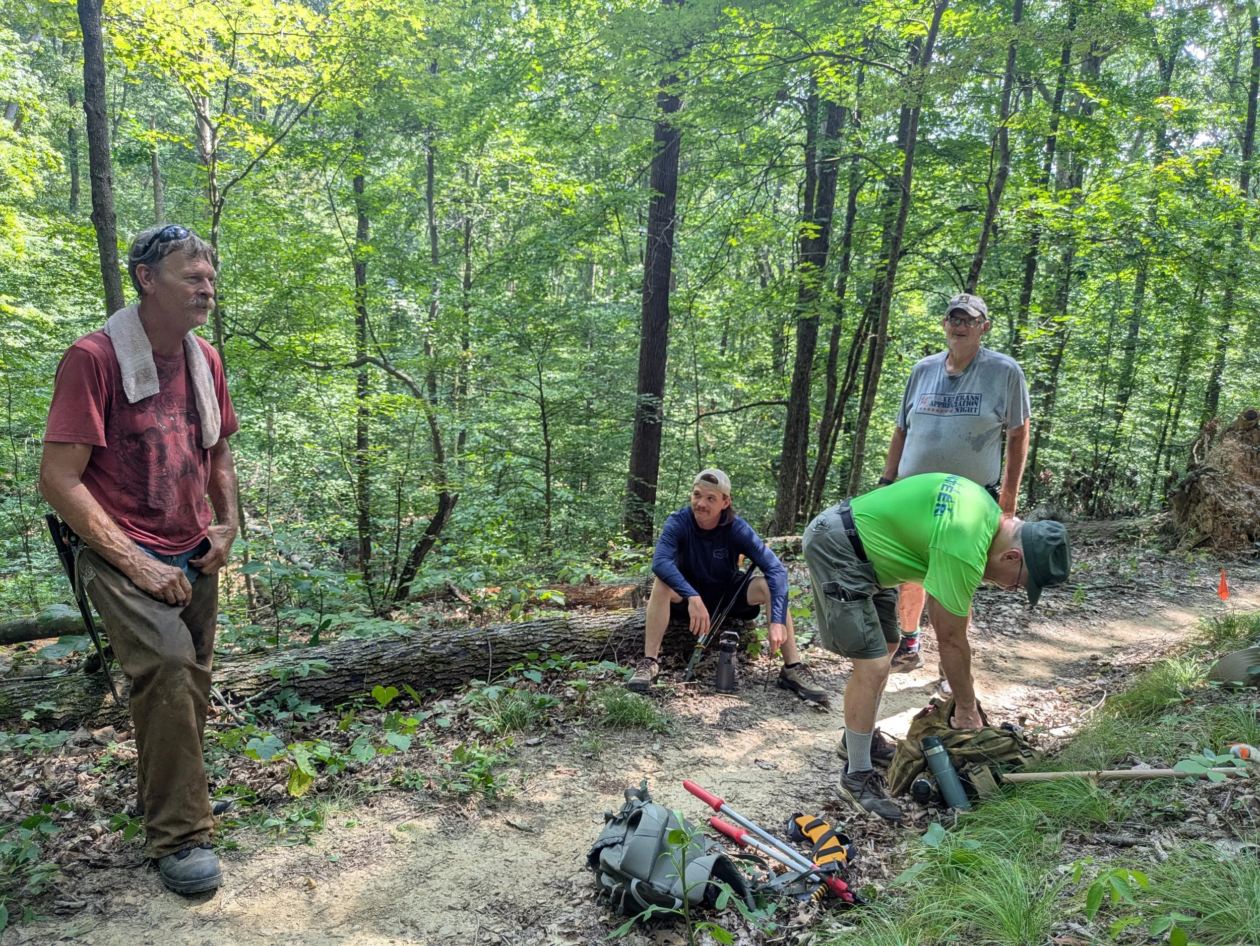 A group of people in a dense green forest, some standing and others bending over, with equipment and backpacks on the ground, possibly on a hiking or outdoor adventure.